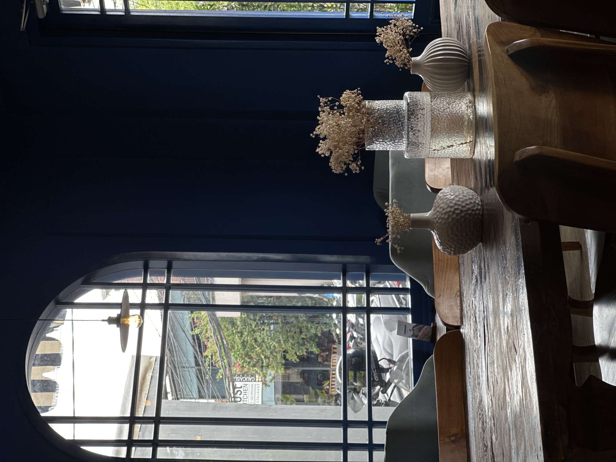 The image shows an indoor scene, likely a cafe or restaurant in North Kuta, Indonesia, as evidenced by signage visible through the windows. A table made of light-colored wood is prominent, with two white vases and one clear, textured vase containing dried flowers arranged on its surface. Part of a wooden chair's back and armrest are visible on the right side of the frame, indicating seating at the table.
Outside the arched window, the street features buildings, trees, and parked scooters or motorcycles. Signage in the background shows "CAUCASIAN KITCHEN GEORGIAN RESTAURANT" The atmosphere is bright due to natural light filtering through the window.