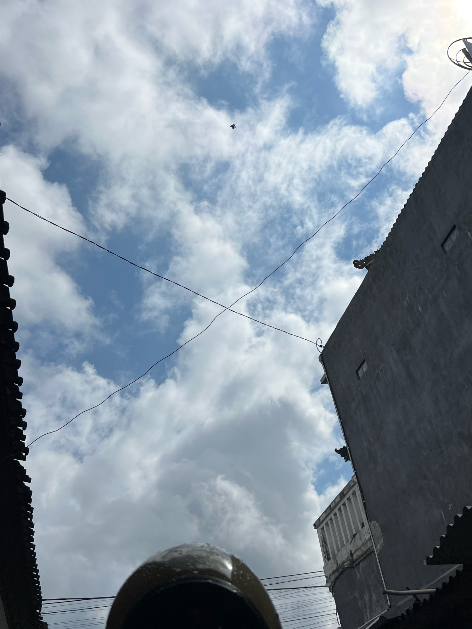 A black object, possibly a drone, is suspended in the sky over Denpasar, Indonesia. The sky is partly cloudy with a blue sky visible between the clouds. The image is framed by the dark, angular roofs of buildings on either side. Thin black wires crisscross the sky, casting faint shadows. A dark grey building with two small rectangular windows occupies the right side of the frame, its roofline visible against the clouds. The image is taken from a low angle, looking upwards.