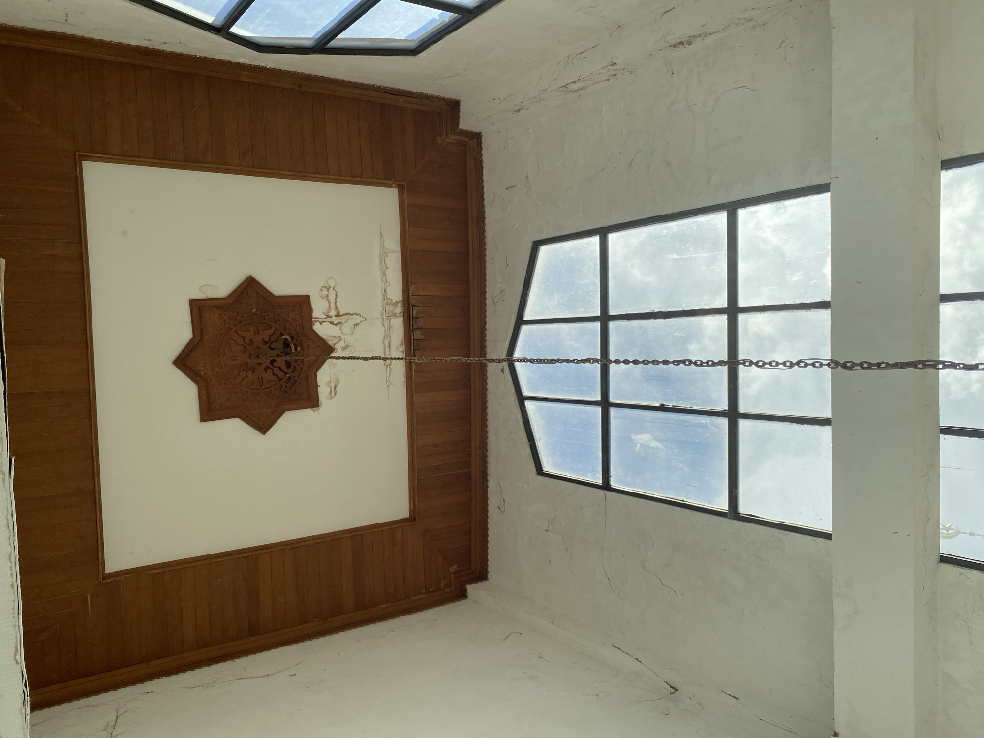 The image shows the interior of a building in Banda Aceh, Indonesia, with a focus on the ceiling and windows. The ceiling features a wooden trim surrounding a white central area with a decorative wooden star-shaped ornament. A metal chain hangs from the ornament, descending towards the viewer. Two large, multi-paned windows with dark frames are visible on the walls. The windows reflect a cloudy blue sky. The walls are painted white and show signs of wear, including cracks and water stains. The overall impression is of an old or neglected building.