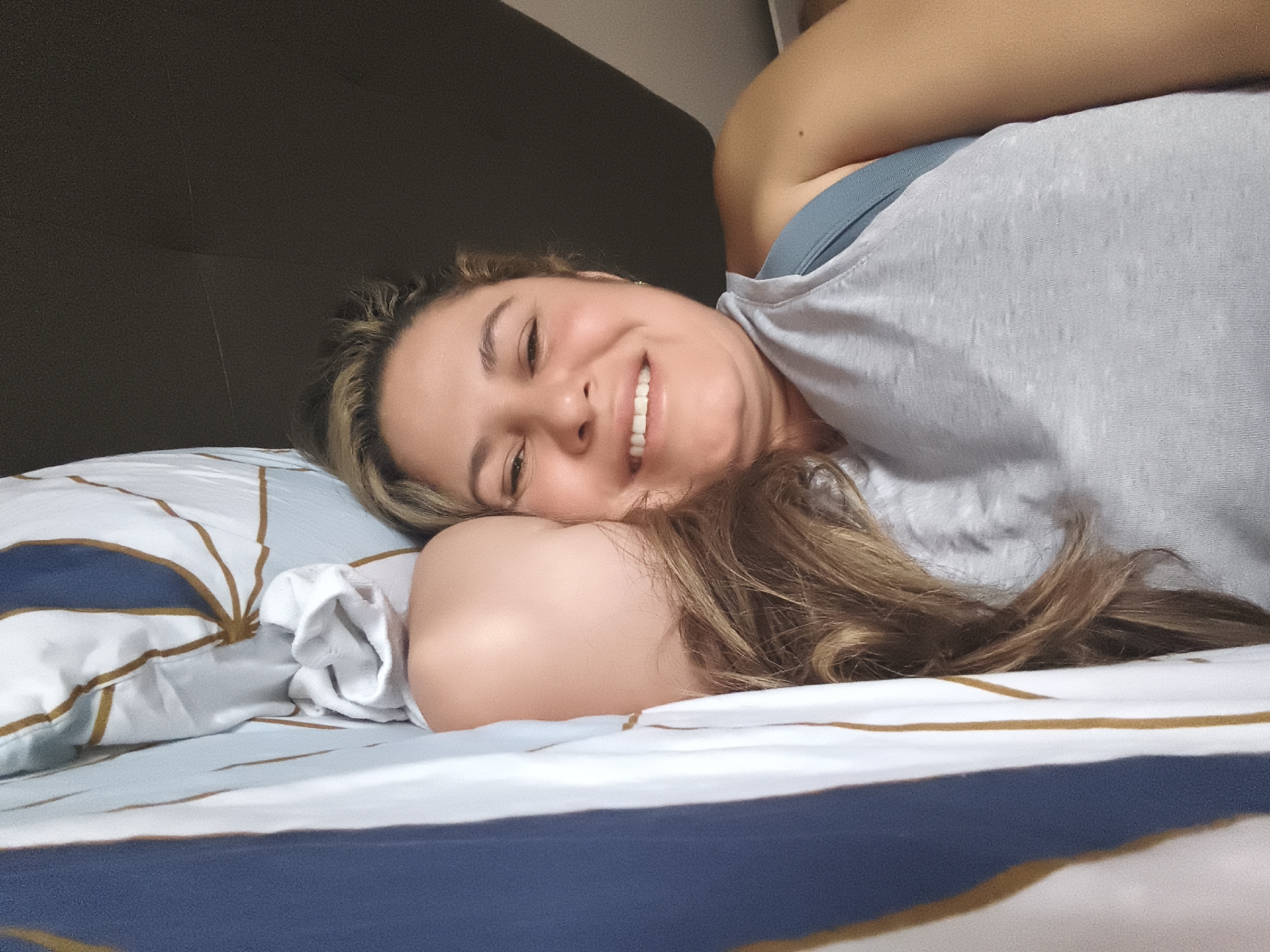 The image shows a woman lying on a bed, with her head resting on a pillow. She is smiling and looking at the camera. The bed appears to be in a bedroom, with a dark headboard behind the woman. The sheets on the bed have a pattern of blue and white stripes with gold accents. The woman is wearing a light blue bra and a grey t-shirt. Her hair is long and brown. The image is taken from a close-up perspective, focusing on the woman's face and upper body. There is no visible indication of the location being in Vitoria-Gasteiz, Spain.