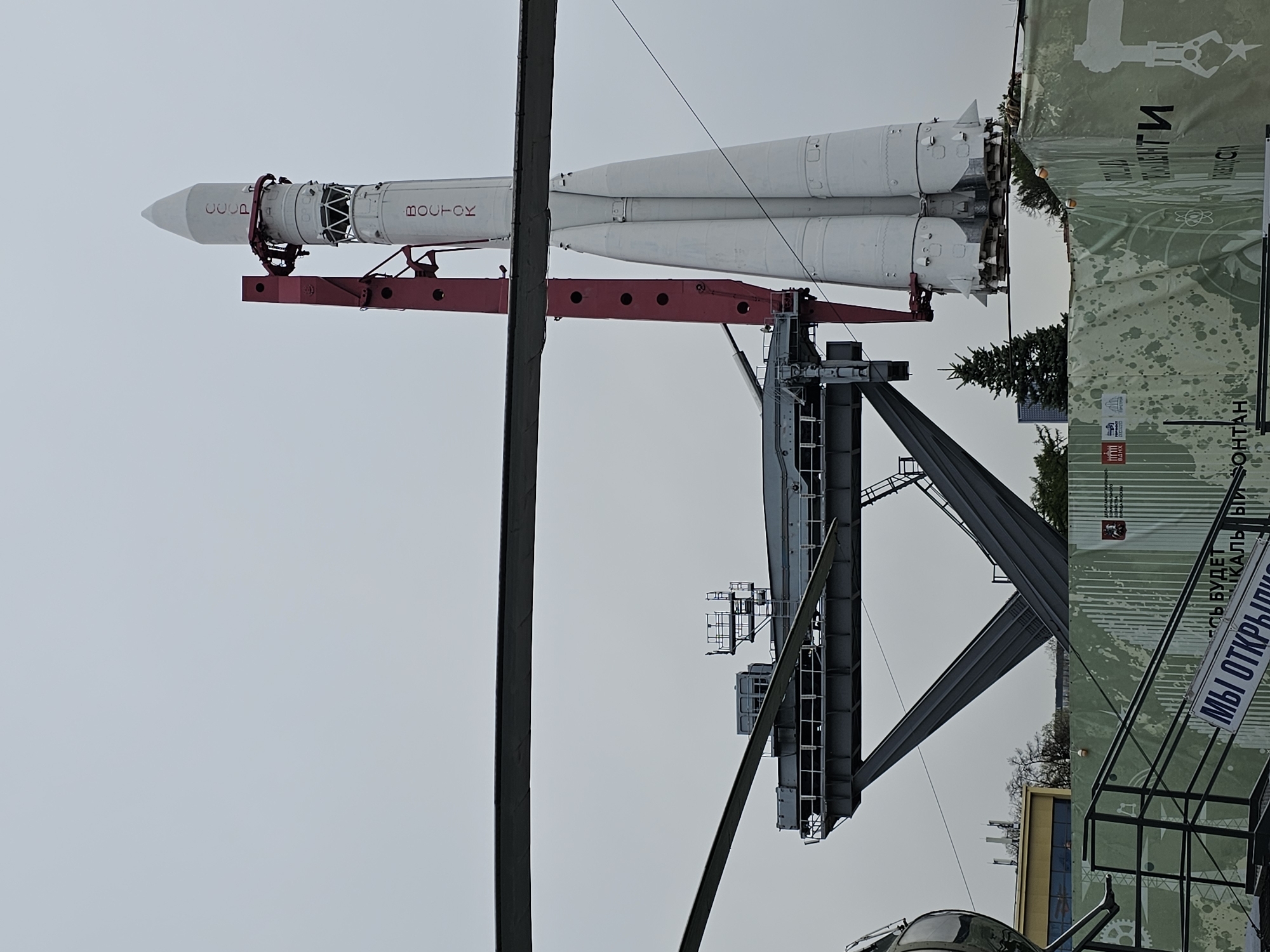 The image shows a Vostok rocket on display at the Exhibition of Achievements of National Economy (VDNKh) in Moscow, Russia. The rocket is white with red lettering and Cyrillic text, mounted on a red support structure. A launch tower-like structure is adjacent to the rocket. Other metallic scaffolding and architectural elements are visible. A banner with Cyrillic writing stands nearby. The sky is overcast and light gray.