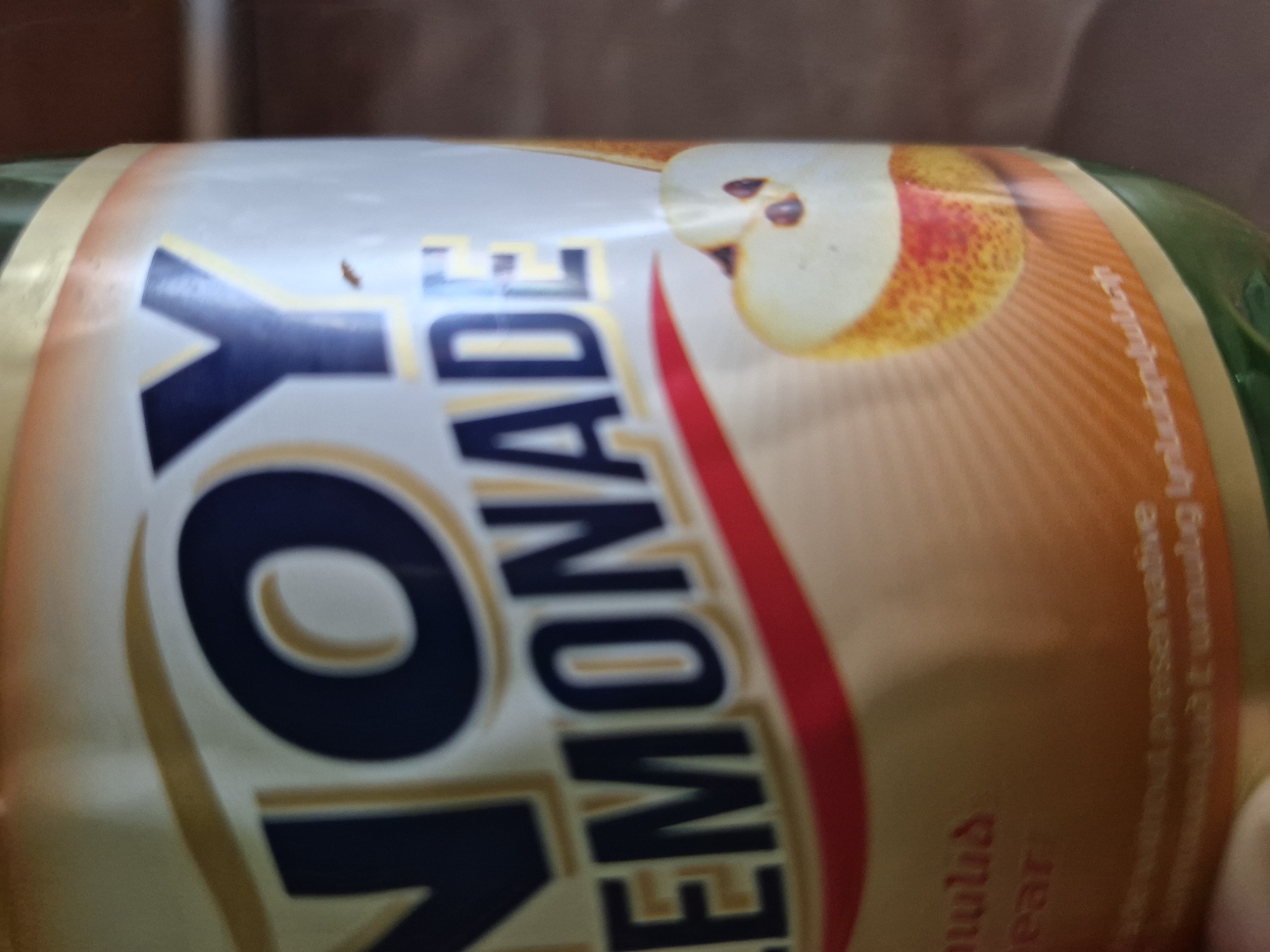 A close-up, slightly angled shot captures the label of a "NOY LEMONADE" bottle, likely in an indoor setting in Yerevan, Armenia. The bottle is positioned horizontally. The prominent text "NOY" is in large, dark blue letters with a gold outline, followed by "LEMONADE" in a similar style. The label features an illustration of a sliced pear, signifying the flavor. A small, dark brown speck, possibly a tiny insect or particle, is visible on the letter "Y" of "NOY."
To the right of the main text, partially visible English words include "pear" and "preserve" (likely part of "without preservative"). Armenian text is also present on the label; "տանձ" (pear) is visible near the pear illustration, and "առանց պահածոների" (without preservatives) is partially seen further to the right. The background is blurred, suggesting a shallow depth of field. The lighting is soft and even, indicating an indoor environment.