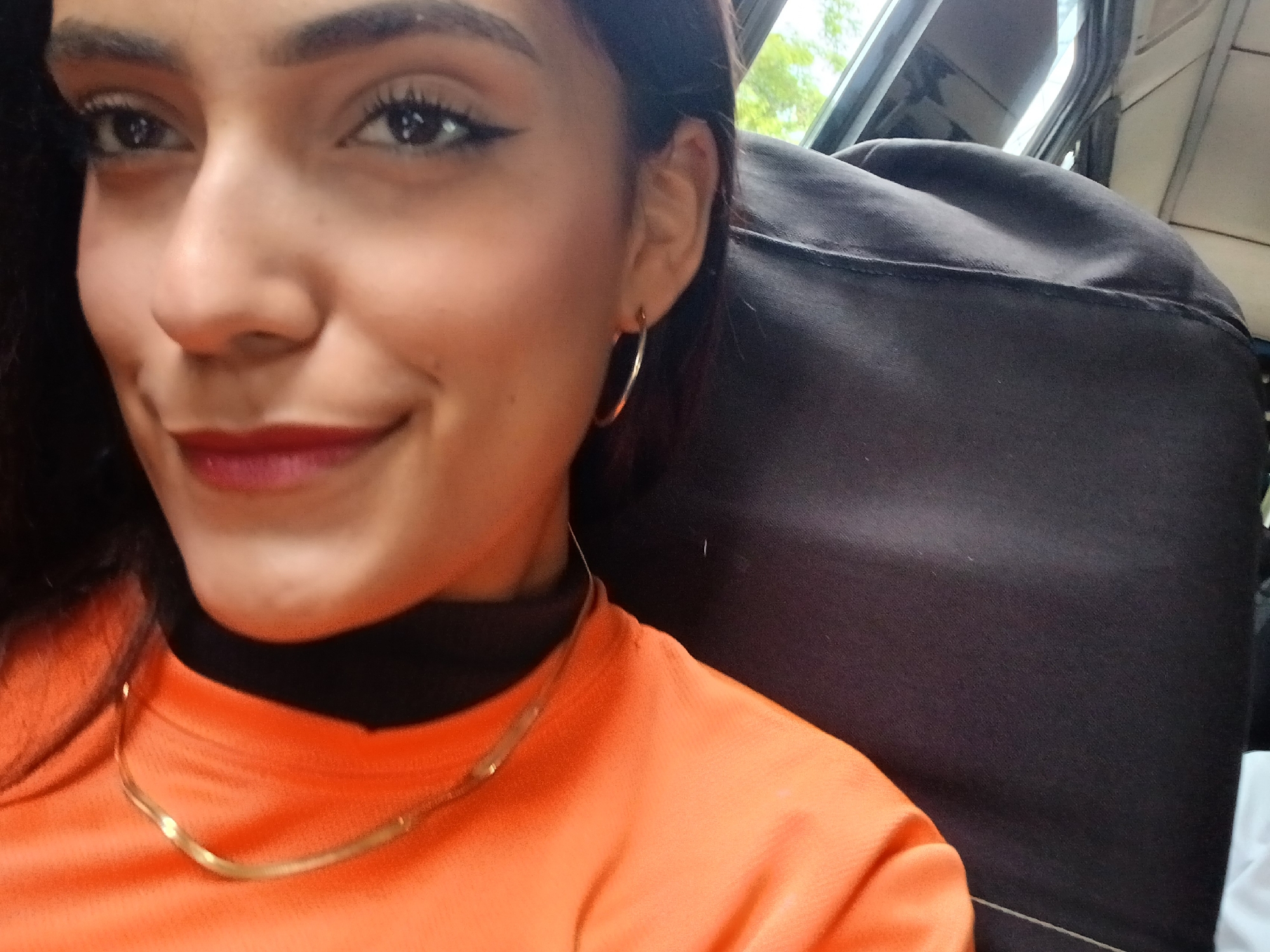 A medium close-up, outdoor, selfie-style shot shows a young woman from the chest up, looking towards the camera. She has dark hair, dark eyes, and is wearing red lipstick. Her left ear is visible, adorned with a thin, gold hoop earring. She is wearing an orange long-sleeved shirt with a black collar and a thin gold necklace. The background consists of the interior of a vehicle, with a dark-colored seat and a window showing greenery outside, suggesting it might be a car or bus. The overall lighting is natural, with some shadows. The location is not identifiable from the visual information provided.