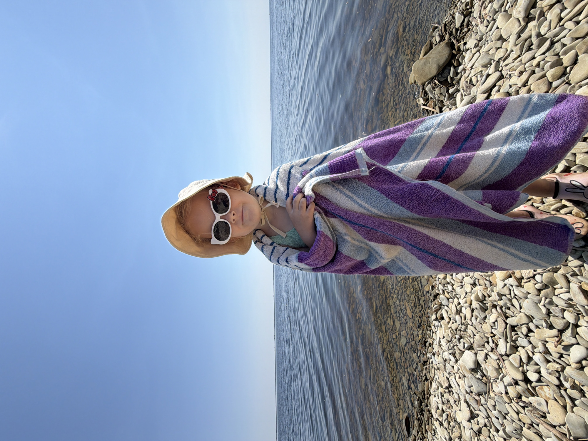 A young girl is wrapped in a striped towel on a rocky beach next to a body of water. She is wearing a sun hat and white sunglasses with red bows. The water is calm and reflects the bright blue sky. The beach is covered in smooth, grey stones. The image appears to be taken on a sunny day, likely in the afternoon given the bright lighting. The girl seems happy and is looking at the camera. There is no visible text in the image. The scene suggests a leisurely day at the beach, possibly after swimming.