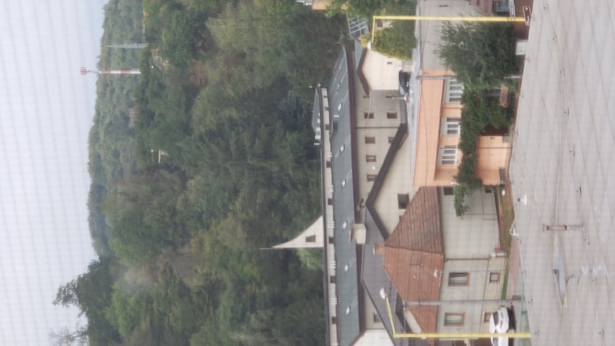 The image captures a view of a residential area in Prešov, Slovakia, with buildings nestled against a tree-covered hillside. In the foreground, a flat, grey rooftop is visible, dotted with numerous metal fixtures. Further back, a light-colored building with a brown, pitched roof is prominent, featuring several windows and a light pole. Adjacent to this is a peach-colored, flat-roofed building with numerous windows and dense green foliage growing over its lower section. Behind these structures, a larger, multi-story building with a dark, sloped roof and skylights is situated. Atop the hillside in the background, a distinctive white, triangular architectural element with a small window can be seen. The hillside itself is covered with a dense canopy of various shades of green trees. A tall, red and white striped communication tower stands on the ridge of the hill, with another, darker tower to its right. The sky is overcast and a pale grey. A white car is parked on the left side of the image, in front of the light-colored building.