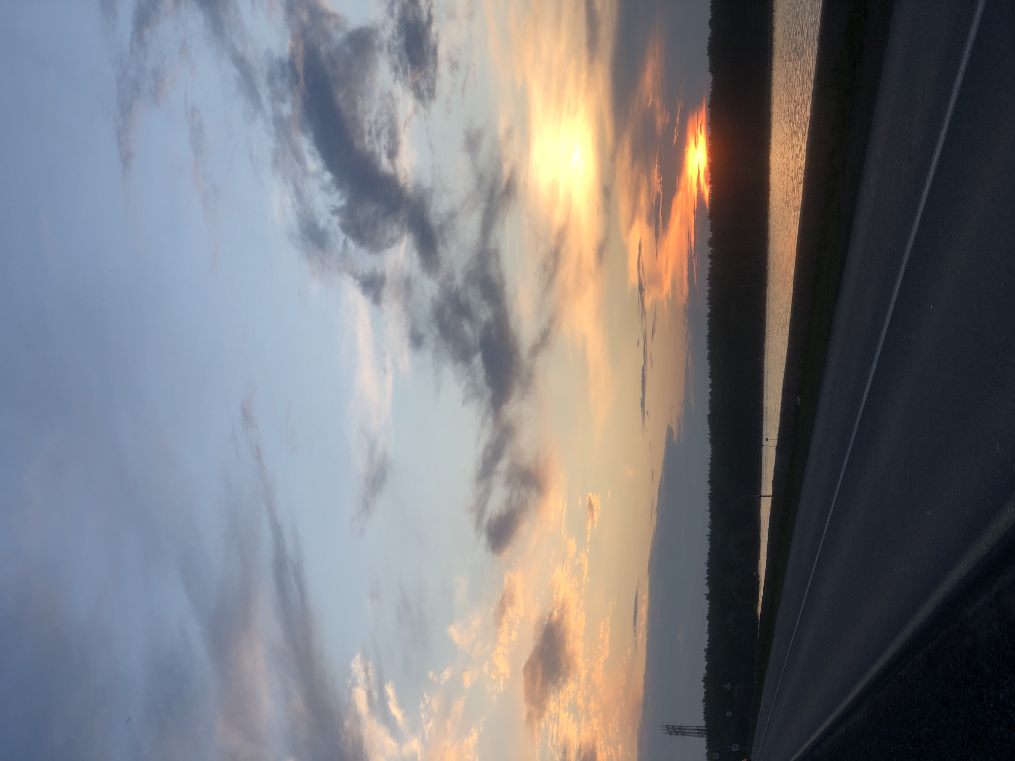 This is a photograph of a sunset over a body of water with a road in the foreground. The sky is filled with a mix of blue, gray, and orange clouds. The sun is a bright yellow orb partially obscured by clouds, casting a warm glow on the water. A dark line of trees runs along the edge of the water. To the left of the frame, a dark asphalt road with white markings stretches into the distance, bordered by a grassy shoulder. In the far distance, a tall lattice tower is visible. The scene appears to be located in Chaikovsky, Russia, based on the context provided. There are no people or specific actions depicted in the image.