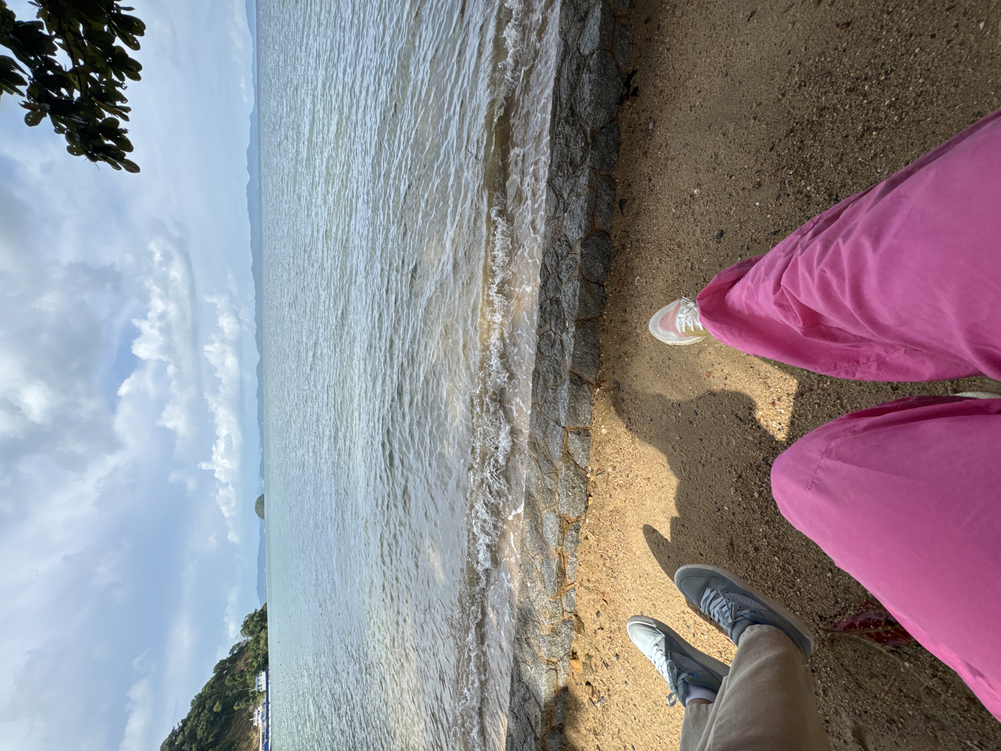 The image shows a close-up view of two individuals' legs and feet sitting on a sandy beach, with the ocean and a distant treed island in the background. The person on the left is wearing light-colored pants and gray sneakers. The person on the right is wearing bright pink pants and white and pink sneakers. The beach is made of sand and small pebbles, with a stone retaining wall separating it from the water. Gentle waves are lapping at the shore. The sky is partly cloudy, and a tree branch with dark green leaves is visible in the upper right corner of the frame. The scene appears to be a scenic coastal area in Pa Khlok, Thailand, likely a beachside location.