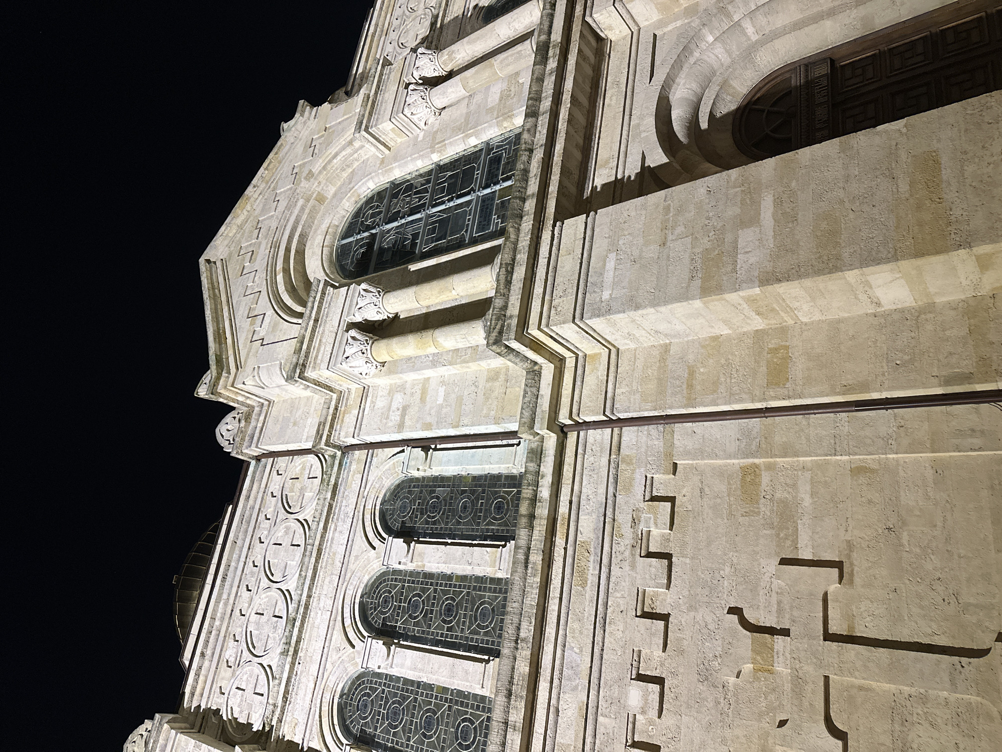 The image shows a portion of the exterior of the Dormition of the Mother of God Cathedral in Varna, Bulgaria, taken at night. The building is constructed of light-colored stone or concrete, featuring intricate architectural details, including rows of rounded windows with ornate geometric designs, columns with decorative capitals, and stepped, geometric patterns. The structure is illuminated, contrasting against the dark night sky. The viewpoint is from below, looking upward at an angle. No people are visible.
