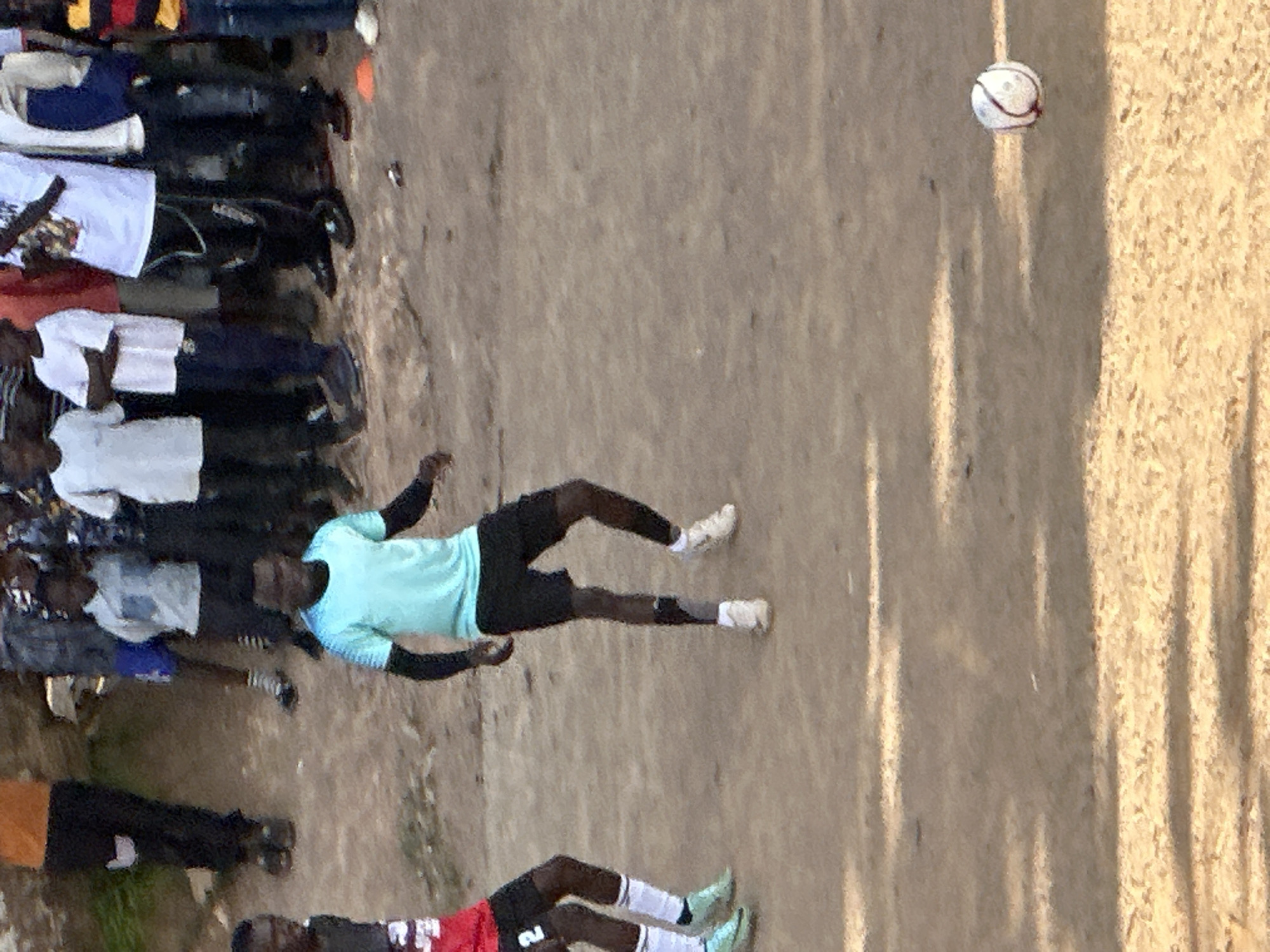 This image captures a moment in an informal football game, likely taking place in Jos, Nigeria, given the context of the prompt. The ground appears to be unpaved, with a sandy, dusty surface indicating a natural field. In the foreground, a young man wearing a light blue t-shirt and black shorts is in motion, mid-stride, with his arms extended. He is wearing white socks and athletic shoes, and a dark strap is visible on his left leg. To his left, another player, partially visible, is wearing a red jersey with the number '2' and black shorts, also in motion. In the background, a group of people, some of whom appear to be spectators or participants in the game, are gathered. They are dressed in various casual attire, including t-shirts, shorts, and pants. A white football with red stitching is visible on the ground in the right side of the frame. The lighting suggests it is daytime, possibly late afternoon, with shadows cast on the ground.