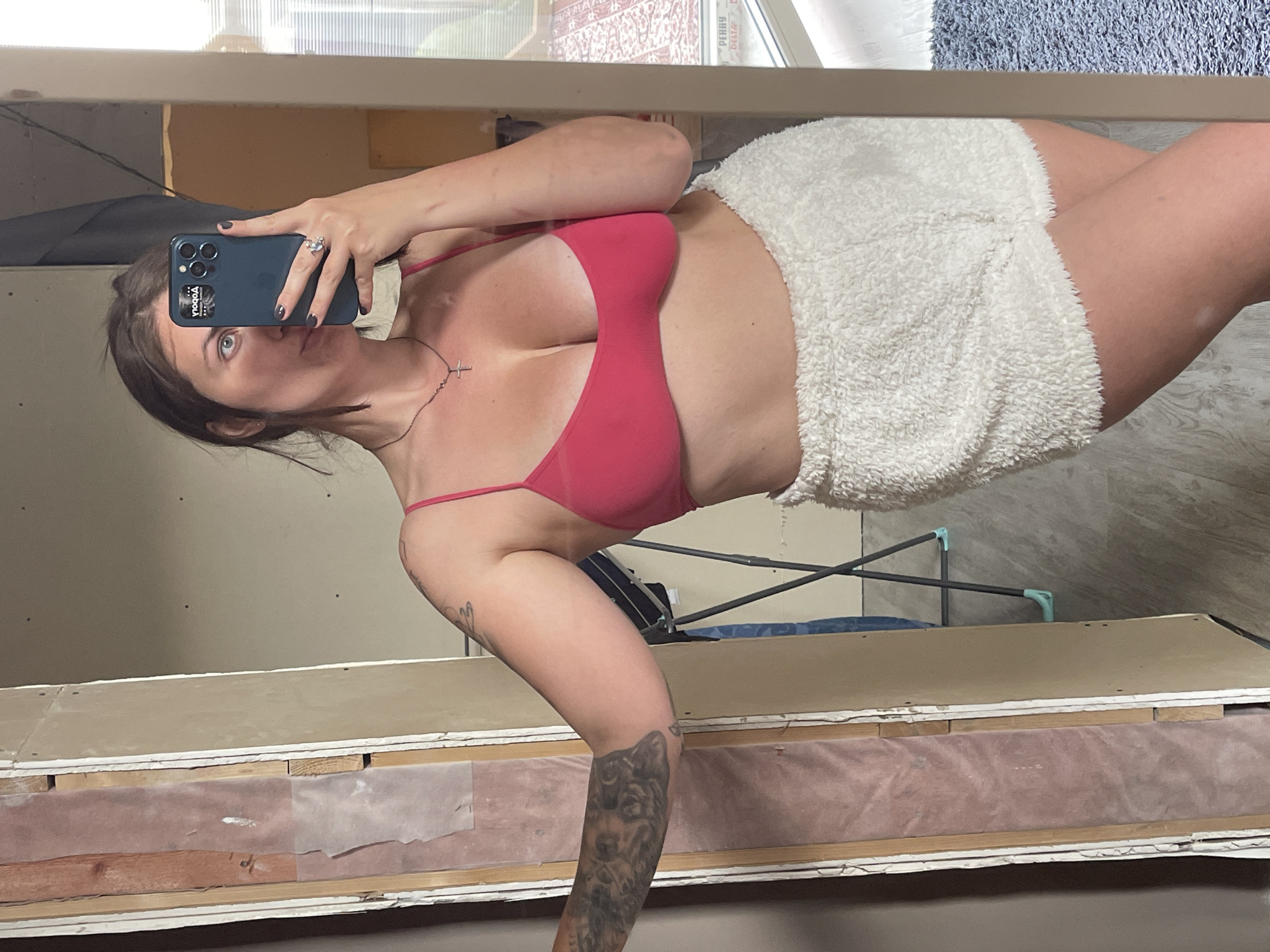 A woman stands in front of a mirror, holding a phone with her left hand to take a selfie. She is wearing a pink top and white, fuzzy shorts. She has a tattoo on her left arm. The reflection in the mirror shows a room under construction or renovation, with exposed walls and wooden beams. A clothes drying rack is visible in the background.
