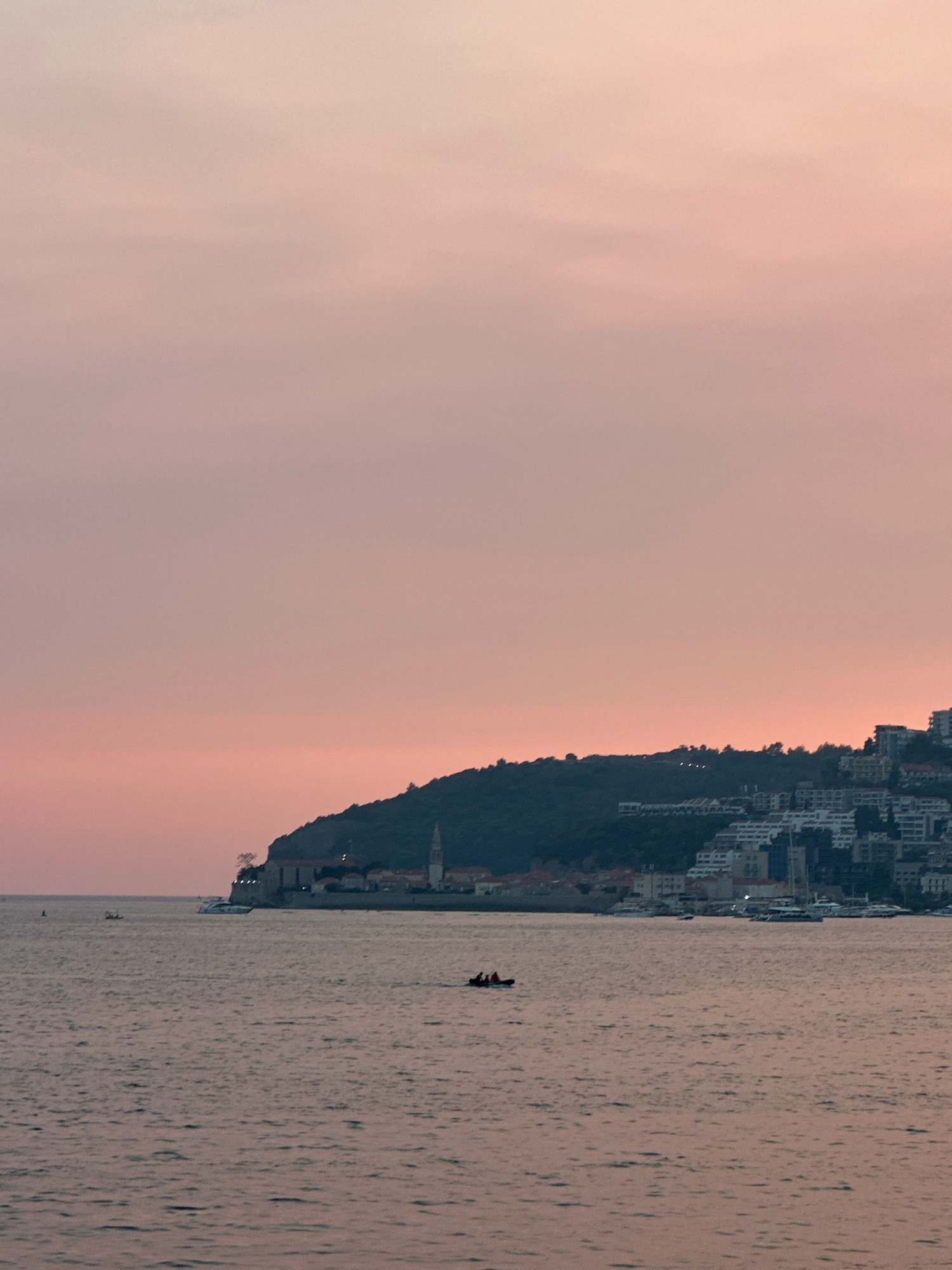 A wide shot captures the tranquil bay of Budva, Montenegro, under a soft, dusky sky tinged with pink and orange. The sea, a muted grey, gently ripples, reflecting the fading light. In the foreground, a small, dark inflatable boat with three occupants navigates the water. Further out, a sleek white yacht is anchored near the shore. The backdrop is dominated by a verdant, tree-covered hill that slopes down to a densely packed cityscape. Buildings, ranging from older structures with terracotta roofs to modern, multi-story apartments with white facades and balconies, are visible along the coastline and climbing the hillside. A prominent church steeple pierces the skyline from the older part of the town, suggesting the historic heart of Budva. The overall impression is one of a peaceful evening in a coastal city.