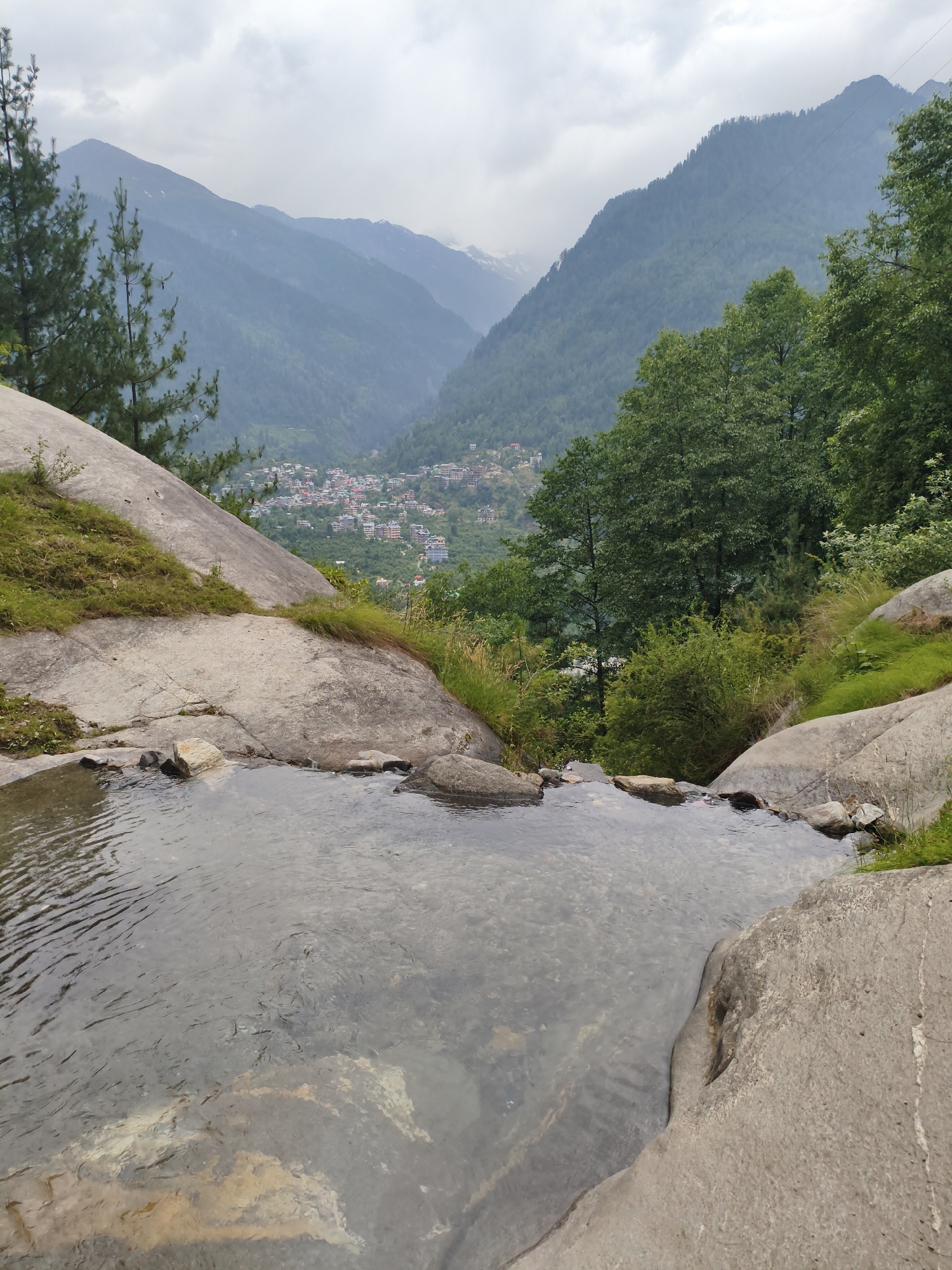 The image captures a scenic view of the valley and the town of Vashisht, India. In the foreground, a shallow pool of clear water with visible rocks and ripples is formed by a gentle cascade. Large, smooth, light-colored boulders frame the pool, with patches of green grass growing on their slopes. Beyond the boulders, the landscape opens up to reveal a lush green valley dotted with houses, indicating the presence of a settlement. The valley is surrounded by majestic, mist-covered mountains, with some snow-capped peaks visible in the distance. The sky is overcast with clouds, casting a soft, diffused light over the entire scene. Tall green trees stand on the right side of the image, adding to the natural beauty of the environment.