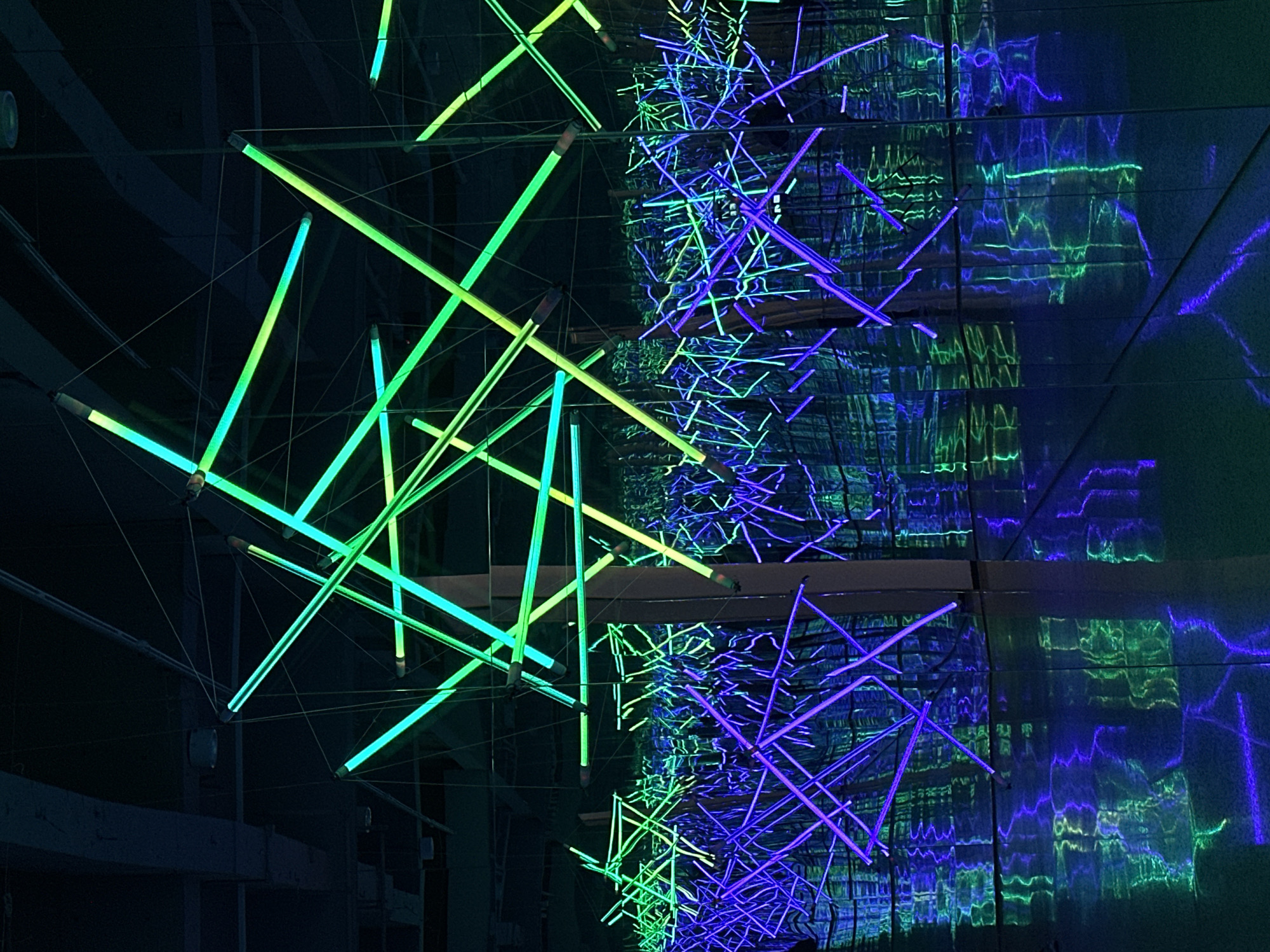 An abstract image shows a dark setting with two clusters of illuminated rods hanging from the ceiling. One cluster on the left is composed of green rods. The other cluster on the right is composed of blue rods. The rods are arranged at various angles and are supported by thin wires connected to the ceiling. The backdrop appears to be reflective, possibly mirrors or polished surfaces, which create a distorted reflection of the rods. The lighting accentuates the rod’s linear shape. The photograph was taken in the city of Banan, China.