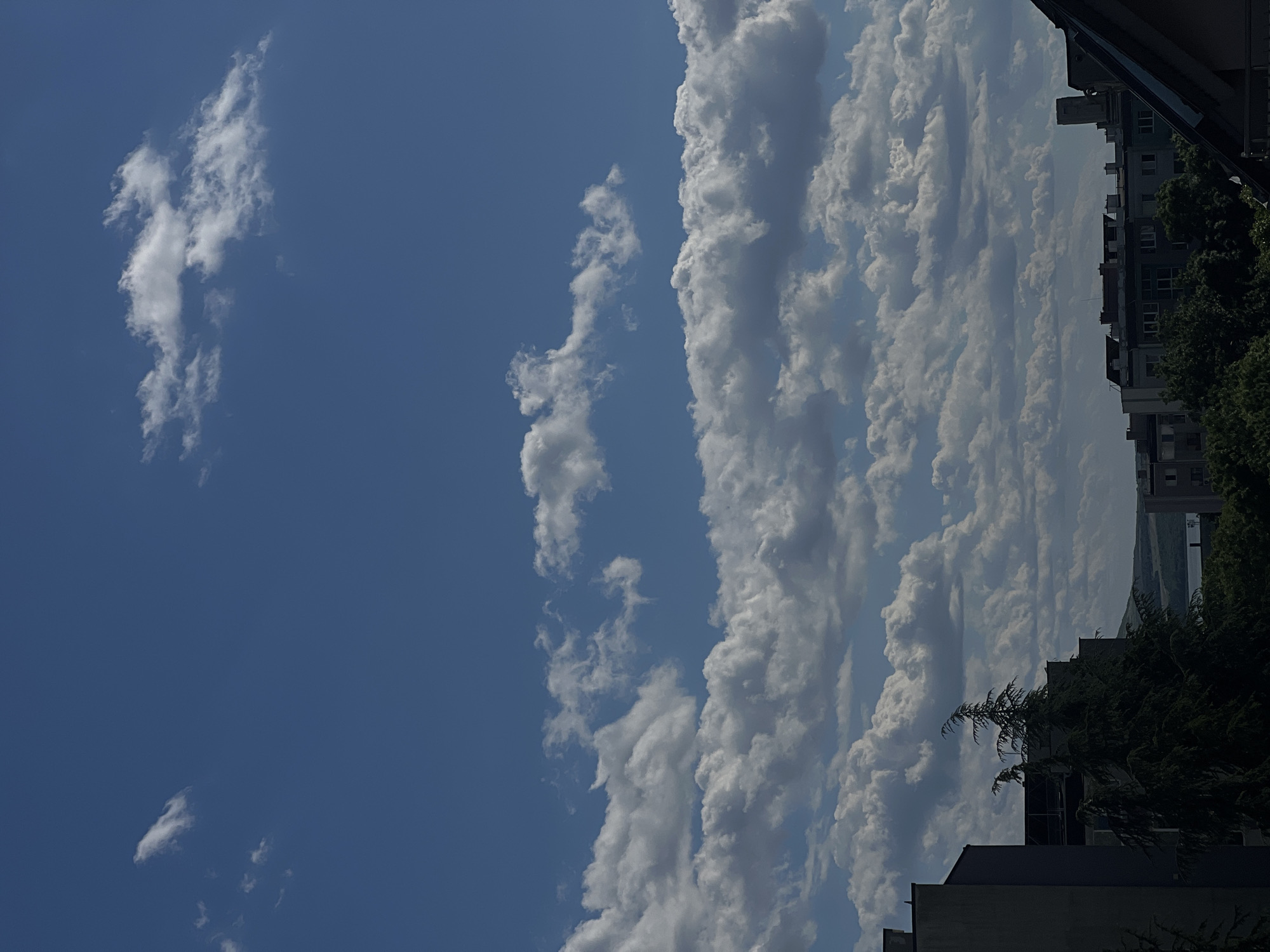 The image presents a daytime view of the sky over an urban area, potentially in Burgas, Bulgaria. The sky features a clear blue section with scattered white clouds, contrasting with a denser, layered cloud formation on the right side. This cloud formation extends vertically and appears to be building, with varying shades of white and gray suggesting depth and potential for precipitation.
In the lower right of the image, the tops of buildings and trees are visible, indicating an urban setting. The buildings are boxy with multiple windows. The presence of greenery, like the trees, suggests the location might be a residential area or a park within the city.