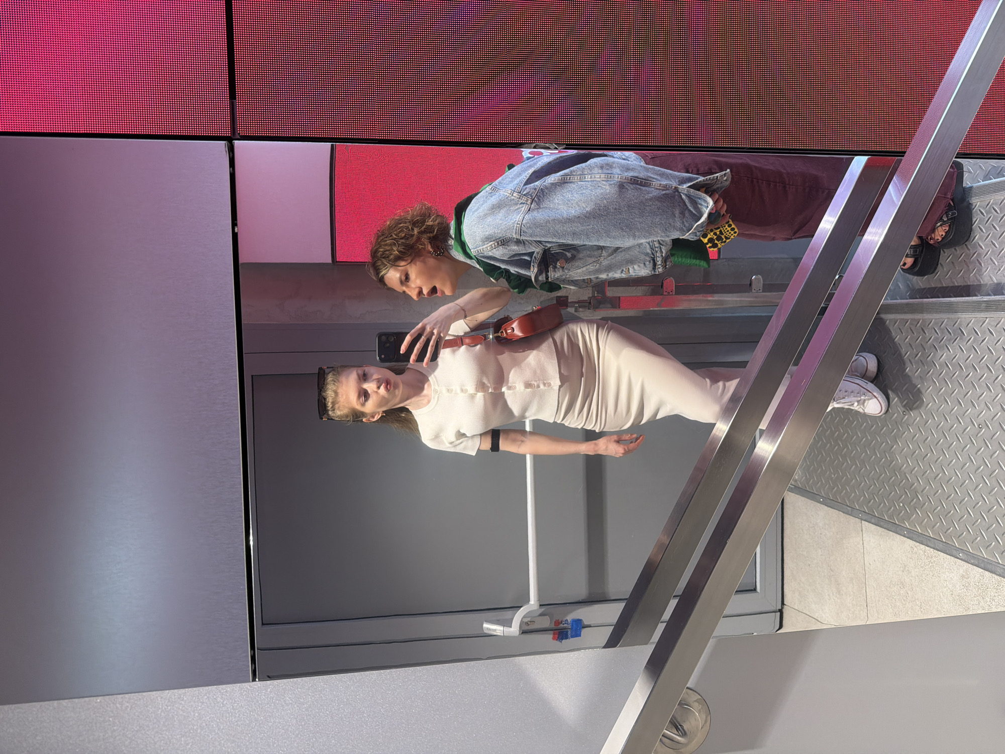 Two women are on a set of stairs. One woman, with blonde hair, is wearing a beige dress and is holding up a phone. The other woman has curly hair and is wearing a blue jean jacket over a green shirt and dark pants. A red handbag is slung over the shoulders of the blonde-haired woman. The stairs are gray metal. There is a large screen with red and gray panels in the background. The photograph was taken in Moscow, Russia.