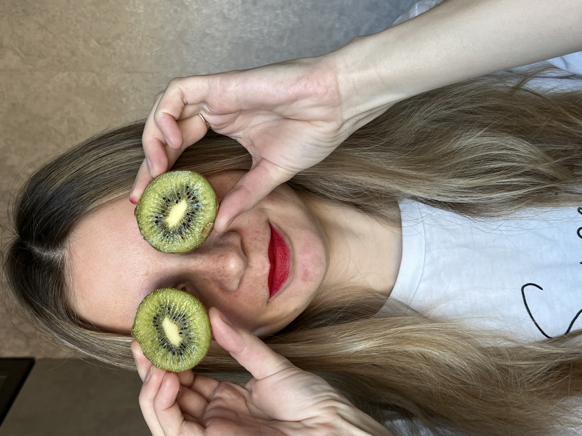 A person is depicted in a playful and relaxed pose, lying down, with long, light brown hair fanned out around their head. They are holding two freshly sliced kiwi halves over their eyes, with their hands visible in the frame. The person is wearing red lipstick, and their skin shows some natural freckles or minor blemishes. A simple, thin ring is worn on the ring finger of one hand. The visible clothing is a white t-shirt with a partial black, curved line design. The background is a plain, light brown or beige surface. The image is taken in Chaikovsky, Russia. No specific time of day or weather conditions are discernible.