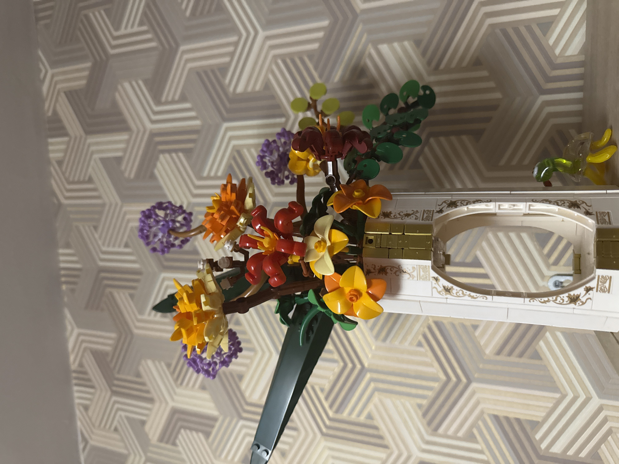The image shows a colorful Lego flower bouquet with various flower types and colors, including purple, orange, yellow, and red. The bouquet is situated within a Lego-constructed architectural element that appears to be part of a building or room, featuring white and gold details, including an arched section. Behind the Lego objects, there is a wall with a geometric pattern of interlocking star-like shapes in shades of beige, gray, and white. The room appears to have a wall painted in a light color to the left of the bouquet. There are no people visible. The location is unknown but cannot be determined to be in Moscow, Russia, without further information.