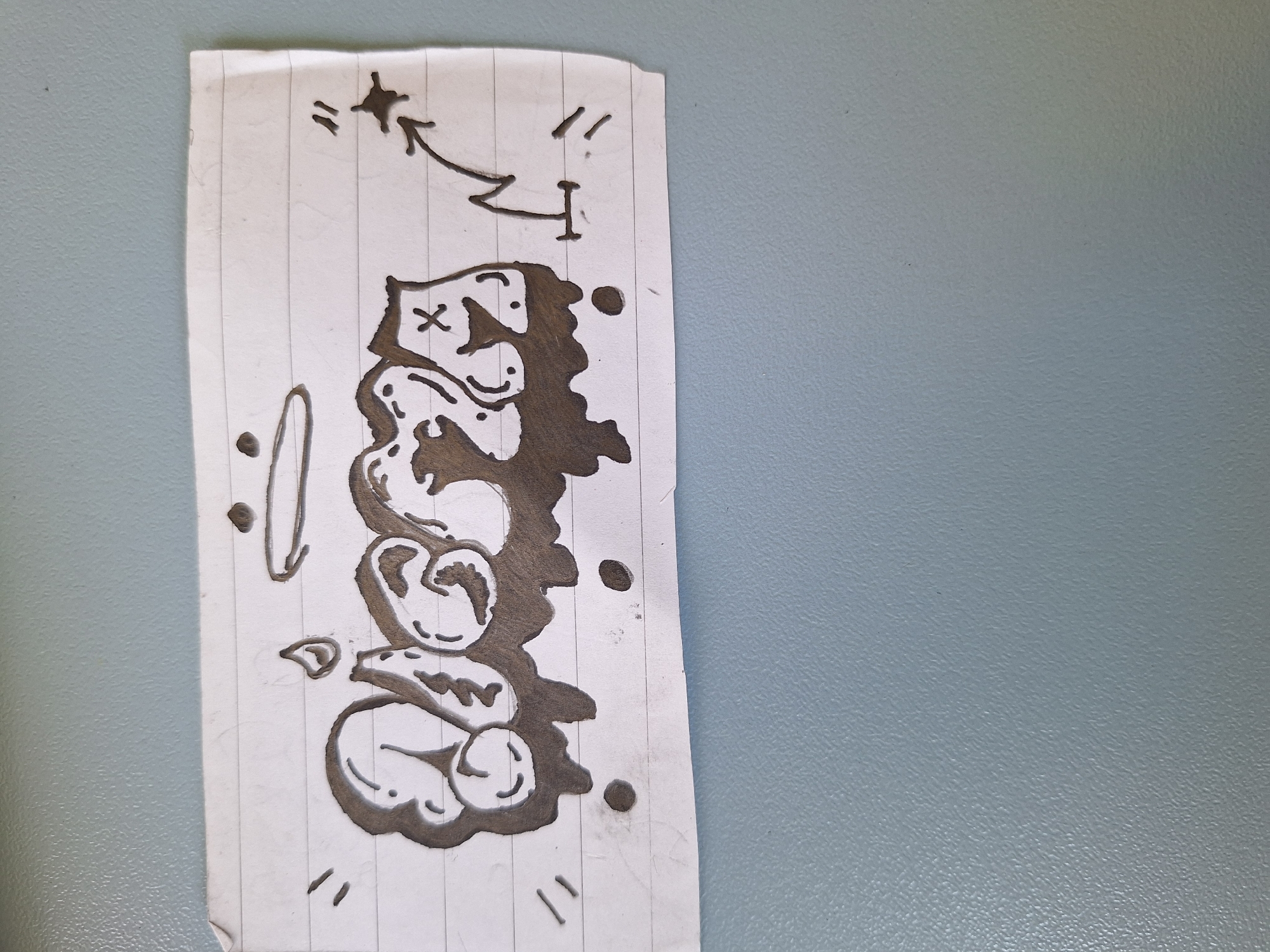 The image is a photograph of a piece of lined paper with a drawing on it. The drawing depicts a word in a graffiti style, with dripping letters. Above the word, there is a halo and a flame. To the right of the word, there is a zig-zag line with an arrow pointing upwards and a star-like shape. The paper is torn on the right edge and has some smudges. The background is a blue, textured surface, likely a wall or cabinet. There are no identifiable landmarks or elements that would indicate a specific location in Varna, Bulgaria. The drawing appears to be a creative expression rather than a depiction of a real-world scene.