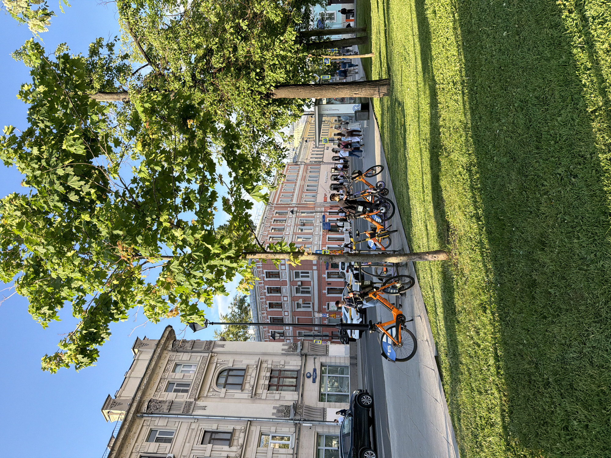 The image depicts a street scene in Moscow, Russia. On the left is a multi-story building with ornate facade details and windows. Parked along the curb is a dark-colored car. A line of rental bicycles is docked along the sidewalk, adjacent to a green lawn. A group of people is standing and walking on the sidewalk in the background. In the far background, buildings in varying architectural styles are visible. Tall trees line the sidewalk, their foliage partially obscuring the buildings. The sky is blue and clear.