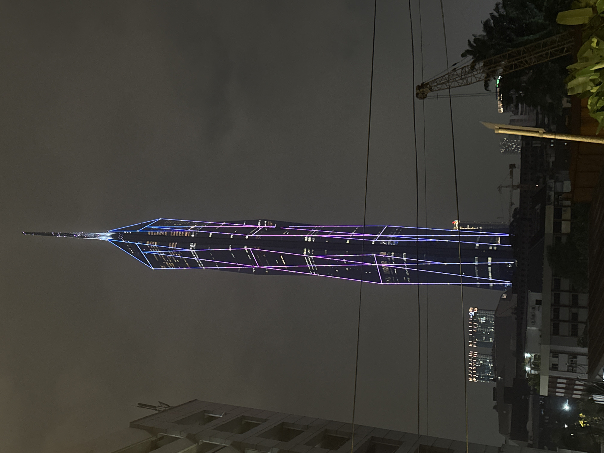 The image presents a striking vertical view of an urban night scene in Kuala Lumpur, Malaysia, dominated by a towering skyscraper. The prominent building, likely Exchange 106, features a sleek, modern design with a tapering spire, illuminated by vibrant purple and blue lights that accentuate its geometric facade. These lights create intricate patterns across the skyscraper's surface, making it stand out against the dark, overcast night sky.
To the right of the skyscraper, a large construction crane with its yellow boom is visible, partially obscured by dark foliage and trees, indicating ongoing development in the area. Utility wires or power lines traverse the upper portion of the frame. In the mid-ground and background, other city buildings can be seen, some with illuminated windows, contributing to the cityscape's nocturnal glow. A streetlight on the right casts a soft light. The time of day is clearly night, evidenced by the dark sky and reliance on artificial illumination. No people are visible, and there is no discernible text within the image. The overall atmosphere conveys a sense of bustling modern urban life and continuous growth.