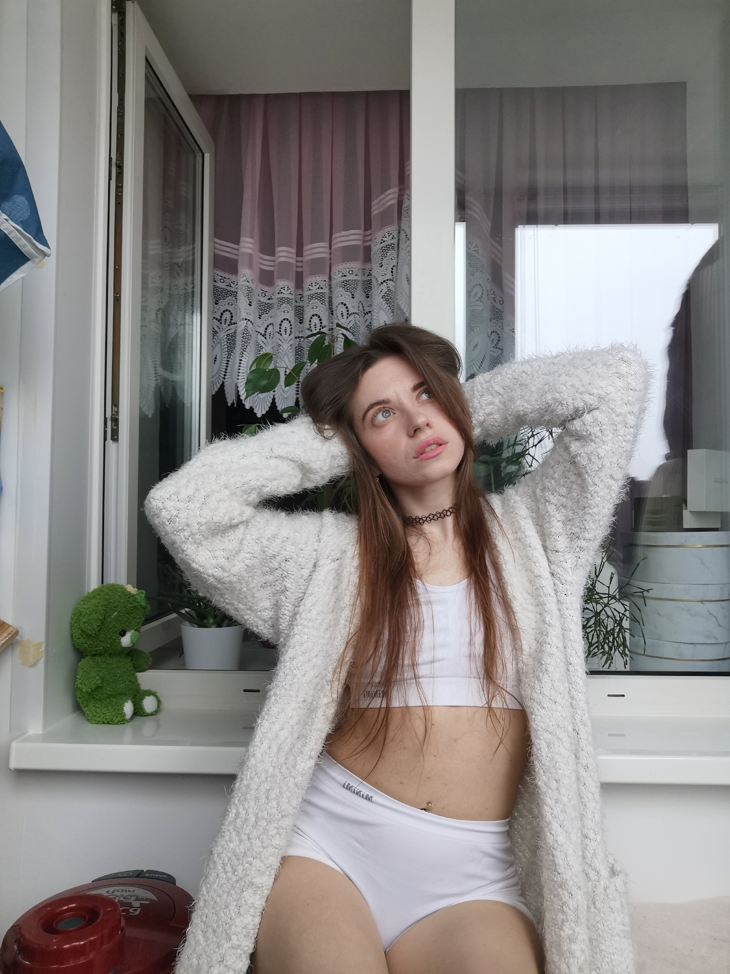 A medium shot depicts a fair-skinned woman wearing a white sports bra, white underwear, and a fuzzy white cardigan. She stands indoors, with her hands behind her head, gazing upwards and slightly to the right. Her long, brown hair is partly loose, with some gathered on top of her head. A black choker adorns her neck.
Behind her, a window is visible, partially obscured by pink curtains with a white lace overlay. Another window shows a cloudy sky and indistinct cityscape. On the window sill to the left, a small green plush toy sits next to a potted plant. To the right, decorative boxes and another potted plant are on the sill. A red vacuum cleaner is partially visible in the lower left corner. The location is in the city of Unknown, Unknown.