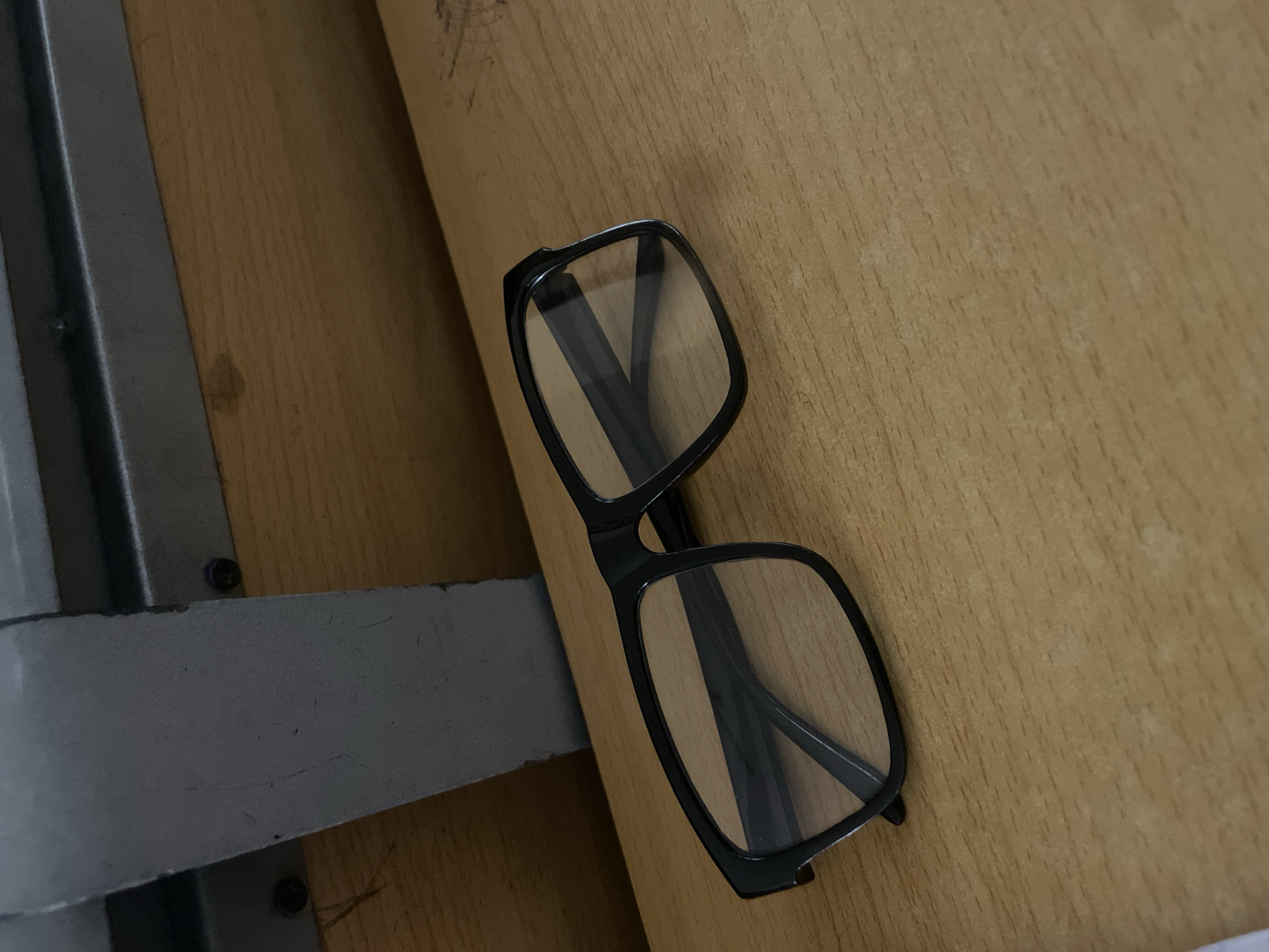 A pair of black-rimmed eyeglasses with clear lenses rests on a light brown wooden surface. The surface appears to be a desk or tabletop. Behind the eyeglasses, a grey metal structure with screws is visible, attached to a lighter brown material that could be wood or laminate. The overall scene suggests an indoor environment, potentially a classroom or office. There is no indication of the specific location in Ihiagwa, Nigeria.