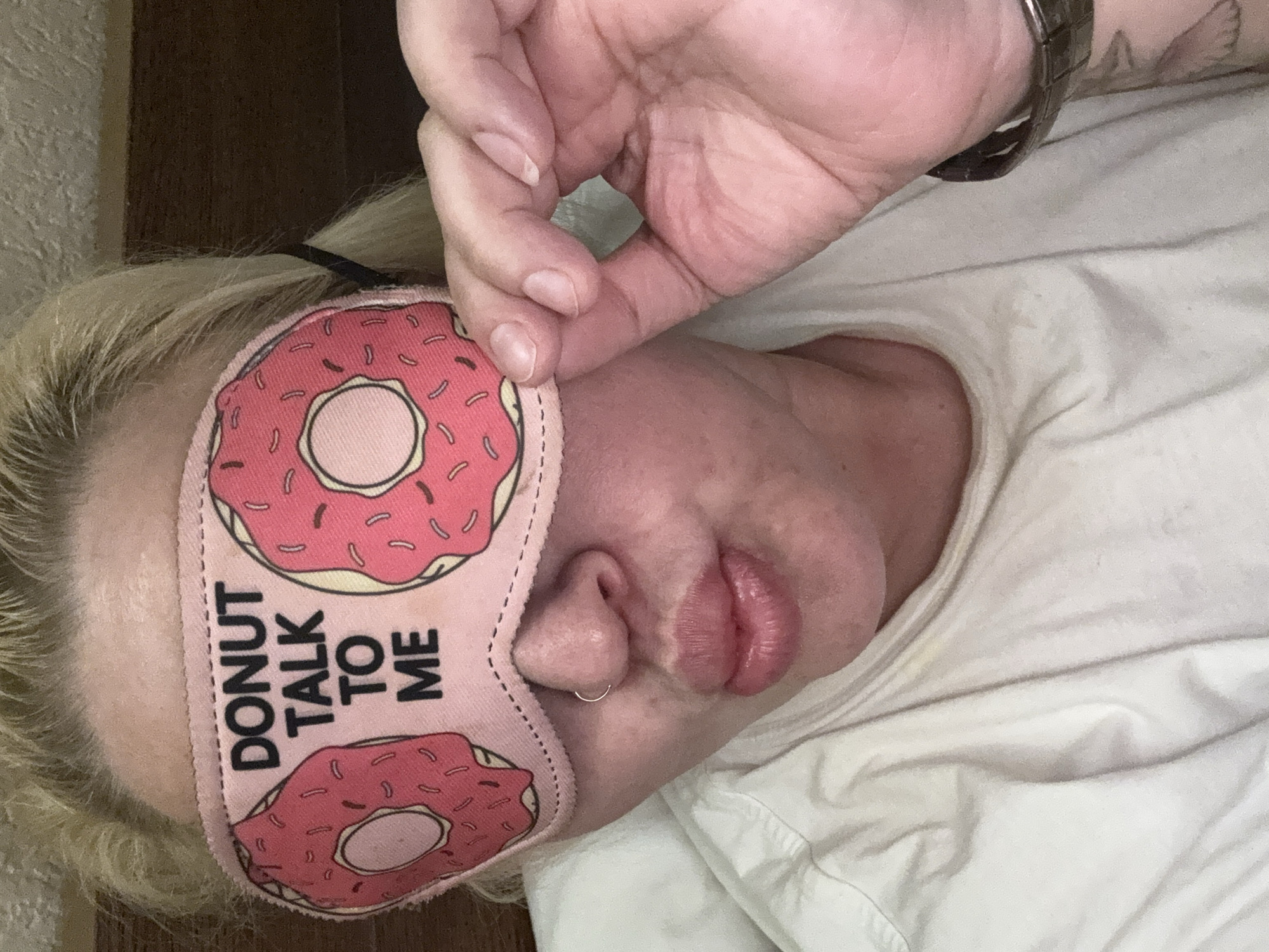 In this close-up, indoor photograph, a person with blonde hair is wearing a pink sleep mask with cartoon donuts and the text "DONUT TALK TO ME" printed on it. The person's eyes are covered by the mask, and their mouth is puckered. A silver nose ring is visible. The person is wearing a white t-shirt and has their right hand raised to adjust the sleep mask. A watch is on their left wrist. The background appears to be a bed with a wooden headboard. The image is taken from a selfie perspective. The location is not specified within the image or the provided context, and therefore cannot be determined to be in Batumi, Georgia.