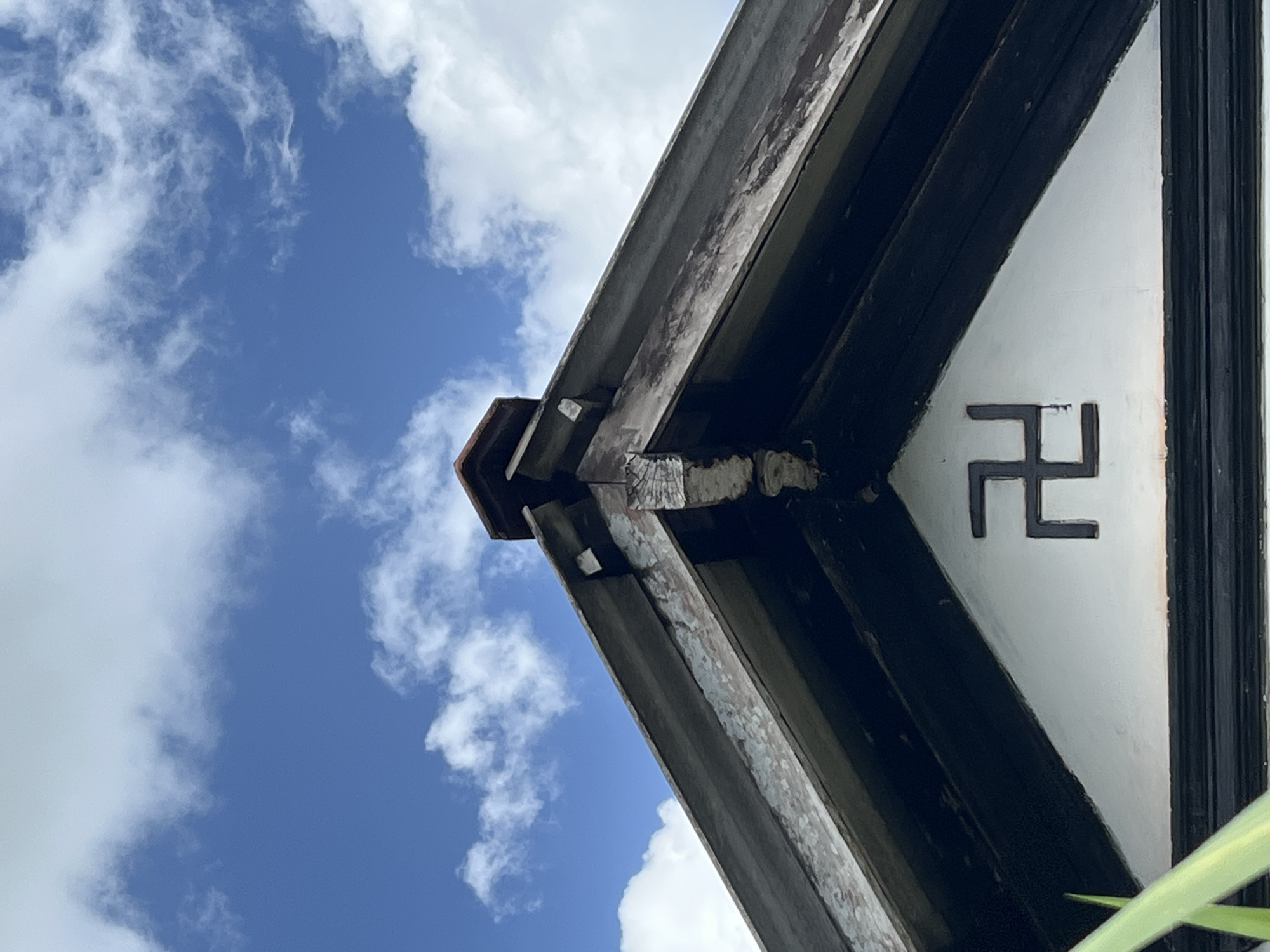 The image presents an upward, angled view of the corner of a building's roof structure set against a bright blue sky filled with scattered white cumulus clouds.
The main subjects are the architectural details of the building's eave or gable. These include several dark, weathered wooden beams that form the framework of the roof overhang. The wood appears aged, with visible texture, some peeling paint, and signs of wear and tear. Beneath these wooden elements, a light-colored, possibly white or off-white, triangular wall section is visible.
On this light-colored wall, a prominent dark symbol is displayed. It is a right-facing (clockwise) swastika, commonly known as the *manji* symbol (卍) in East Asian cultures, where it represents peace, prosperity, and good fortune, often found in Buddhist temples or traditional architecture.
The sky is a clear, vibrant blue, indicating bright, sunny weather during the daytime. A small sliver of green foliage, likely a plant or leaf, is partially visible in the bottom right corner of the frame. No people or specific activities are depicted. The architectural style, particularly the wooden construction and the presence of the *manji* symbol, strongly suggests an East Asian traditional building, possibly a temple or shrine.