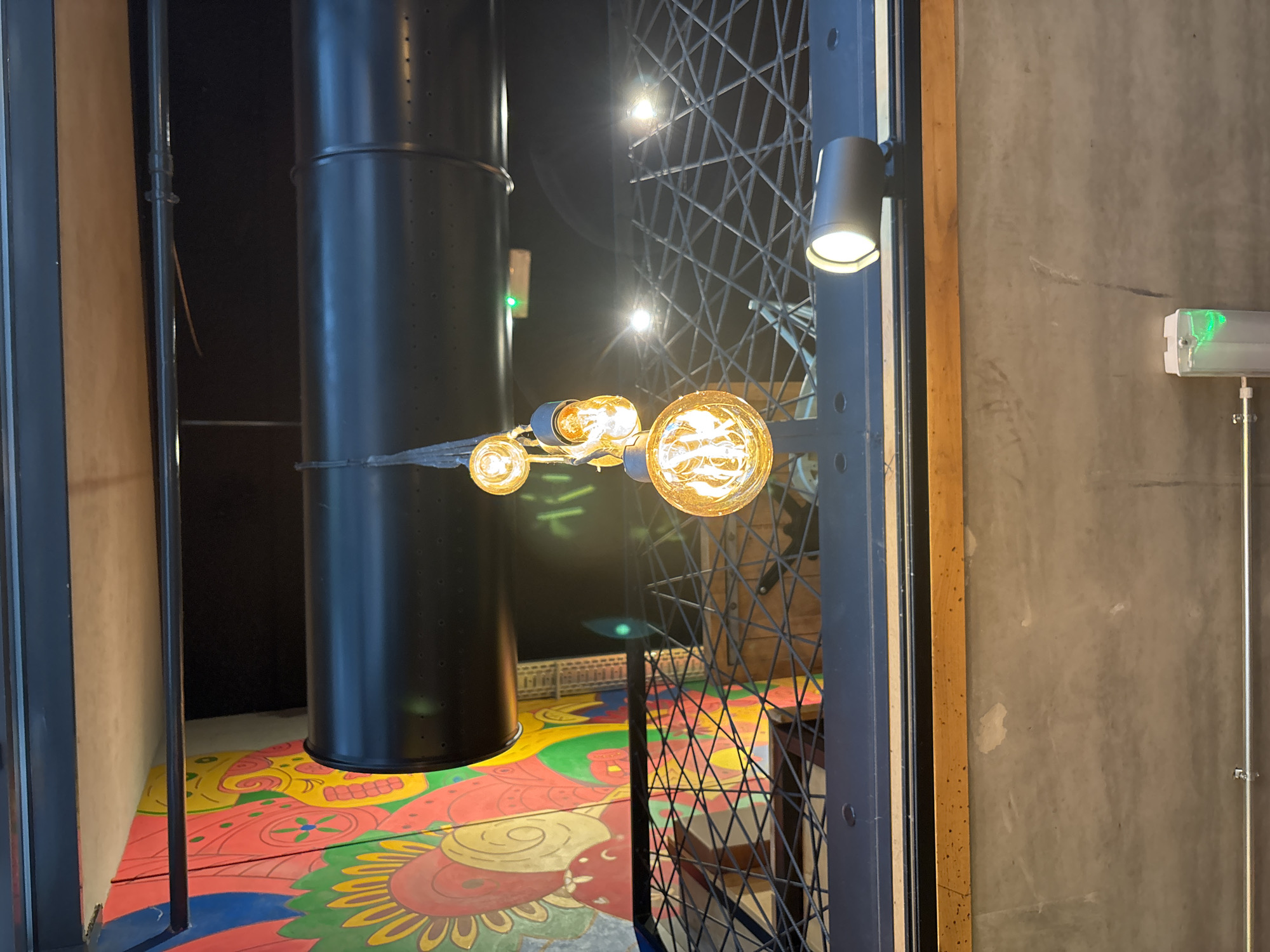 The photograph shows an interior space in Luxembourg City, Luxembourg. A tall, dark metallic structure, possibly a pipe or column, is a prominent element. Decorative light fixtures with exposed bulbs are suspended nearby. The floor is painted with a bright, colorful, floral-inspired pattern. A metal gate or fence with a geometric lattice design is visible. A spotlight and a fluorescent light are mounted on a gray wall adjacent to the gate. The overall setting suggests a mix of industrial and decorative elements in an urban environment.