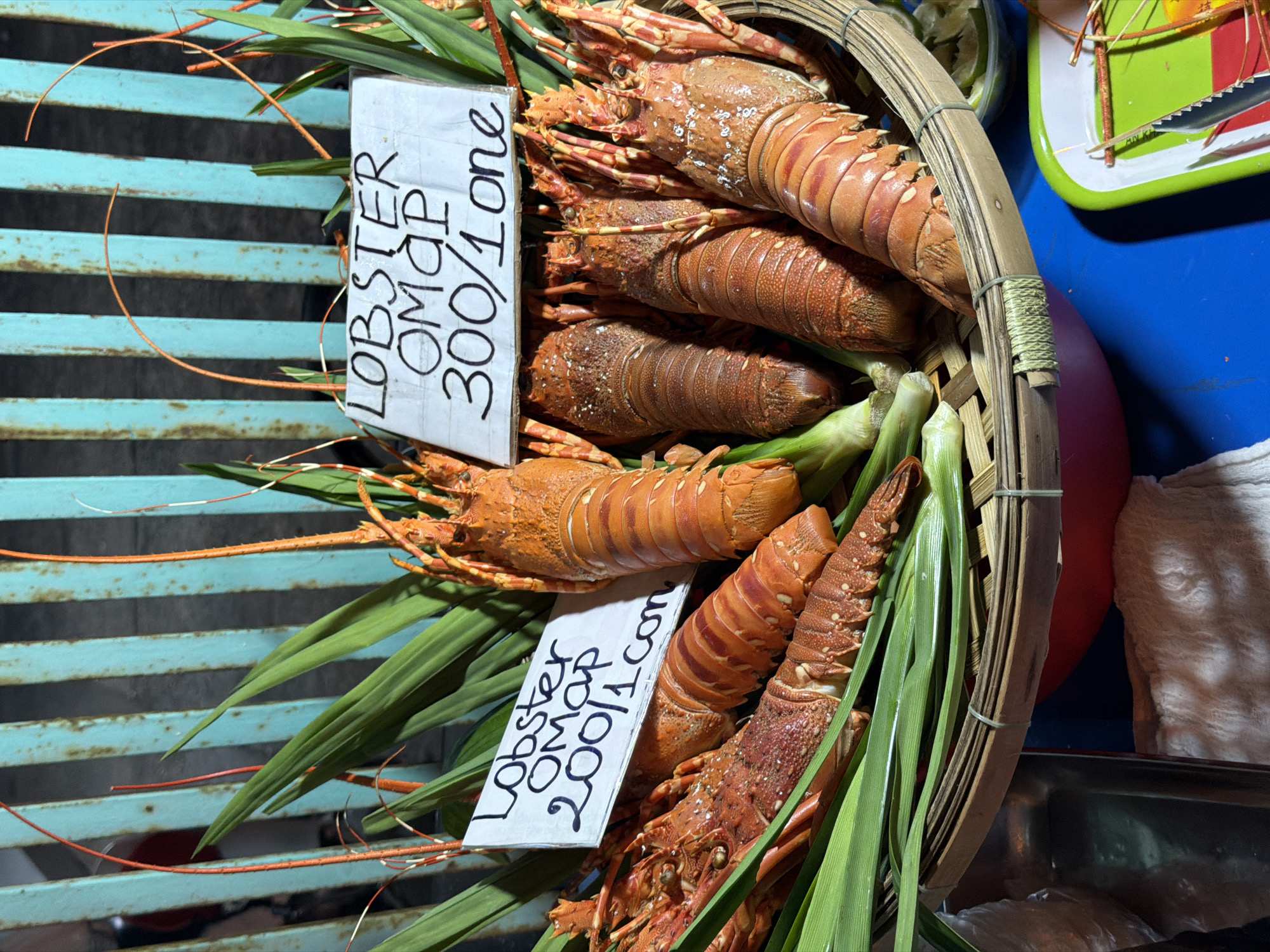 A close-up, eye-level shot captures a selection of lobsters presented in a woven basket. The lobsters, primarily orange and red, are arranged with green leafy vegetables and topped with two handwritten signs. One sign reads "Lobster OMAP 300/1 one". Another sign, partially obscured, states "Lobster OMAP 200/1 con". The background features a teal-colored metal fence with vertical slats and some out-of-focus elements, suggesting an outdoor market or vendor stall. The overall lighting is dim, with some highlights on the lobster shells. The scene is consistent with a street vendor in Nha Trang Ward, Vietnam, selling fresh seafood.