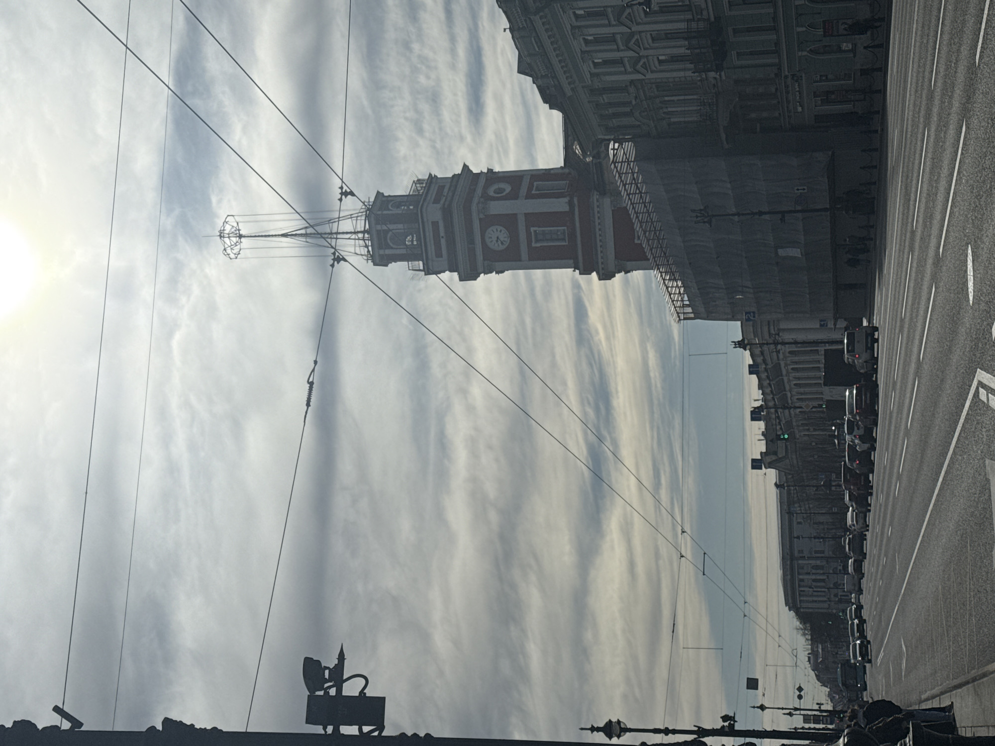 The image captures a broad streetscape in Saint Petersburg, Russia, under a bright, cloudy sky.
The upper and left portions of the frame are dominated by a pale blue sky streaked with elongated white and grey clouds. A luminous, diffused sun is visible on the far left, creating a soft glow. Numerous dark overhead power lines, indicative of trolleybus or tram infrastructure, crisscross the sky, extending throughout the scene.
A distinctive red and white clock tower stands prominently in the mid-ground, slightly to the left of center. The tower features multiple clock faces, one clearly displaying approximately 10:20 or 10:25. Its peak is adorned with decorative metalwork and connects to some of the overhead wires.
To the right of the tower, a row of multi-story, classically styled buildings lines the street, displaying architectural details. Some sections of these buildings, particularly further down the street on the right, appear to be partially obscured by scaffolding or protective coverings.
The lower right part of the image shows a wide, multi-lane asphalt road with clear white lane markings. A line of vehicles, including cars and a small truck, is visible along the road, extending into the distance. Traffic signals can also be seen. The immediate foreground shows dark silhouettes of street furniture or structures, and indistinct figures of pedestrians are visible on the sidewalks along the right edge of the road.
The bright daylight and the position of the sun suggest a morning or early afternoon setting. No legible text is discernible within the image.