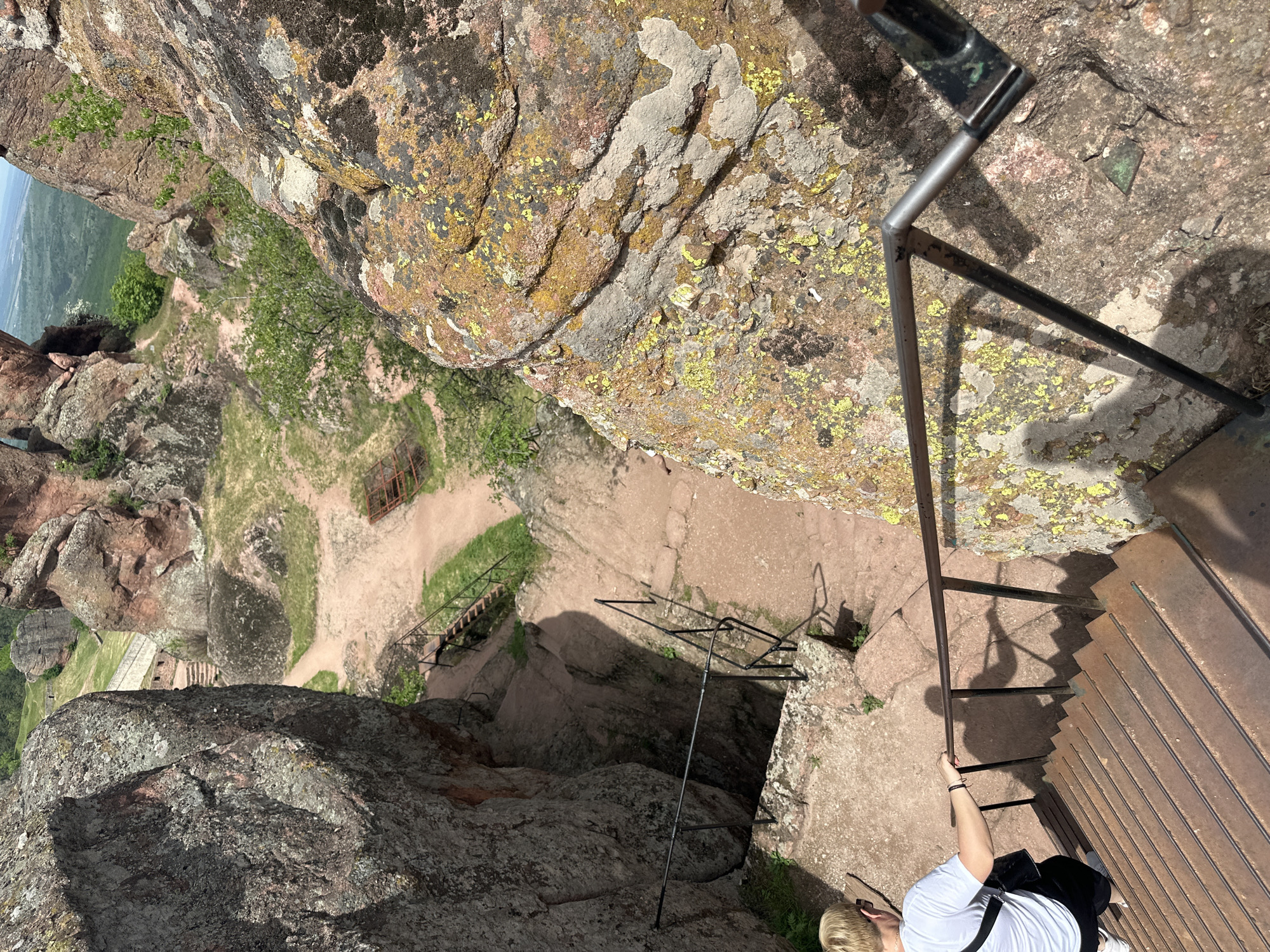 A high-angle shot shows a stairway descending a rocky, steep incline in Belogradchik, Bulgaria. The rock formations are visible on either side of the path. A series of metal railings provide safety. A person dressed in a white shirt and black pants is partially visible at the bottom right corner, holding onto the railing. Vegetation is sparsely scattered throughout the landscape. At the bottom left, one can see a distant valley and greenery.