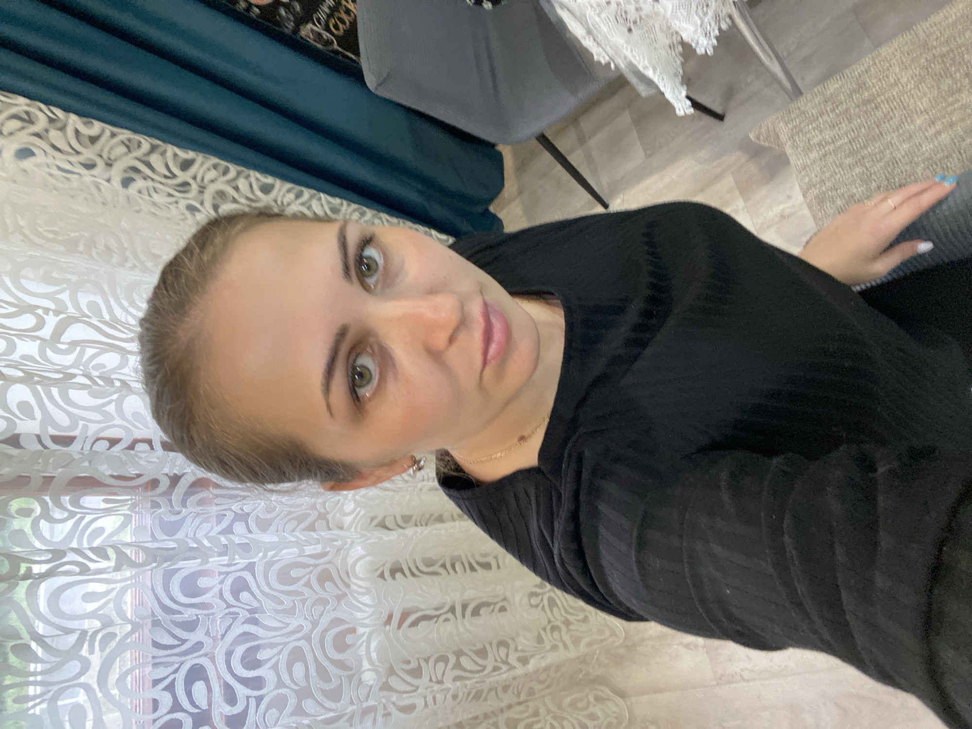 A woman with fair skin, green eyes, and her blonde hair pulled back is looking at the camera. She is wearing a black ribbed long-sleeved top. She is indoors in a room with white patterned curtains, a teal curtain, a grey chair, and a table with a white lace tablecloth. Her hand is visible on a grey upholstered surface. There is no evidence to confirm the location of the image in Марковский, Russia.
