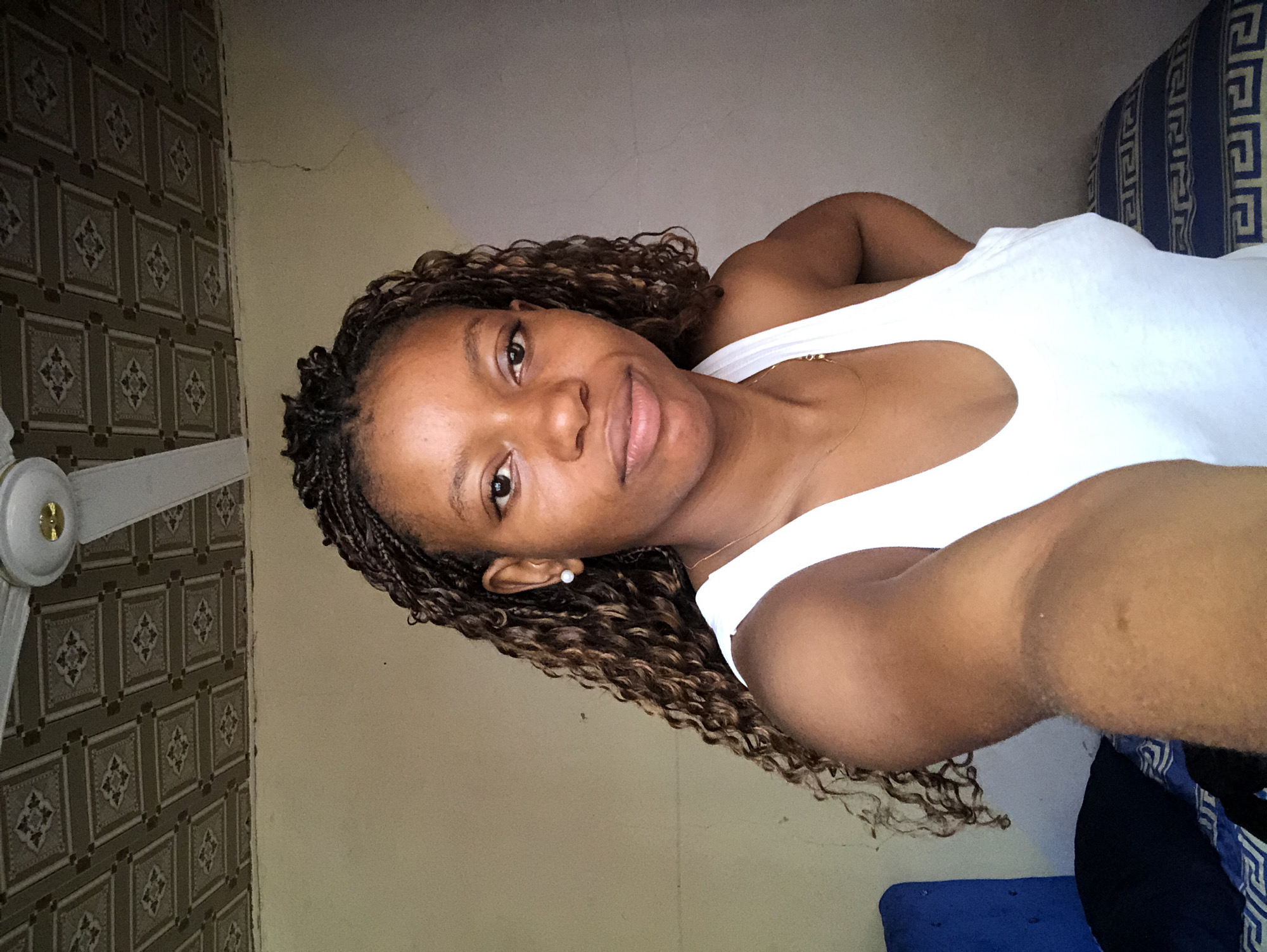 A medium shot depicts a woman with braided hair and a white tank top, seemingly indoors. The setting appears to be a residential space in Kashere, Nigeria, indicated by the presence of specific local architectural elements and interior decor features typical of the region. These include patterned tiles on a wall and a ceiling fan. The woman is positioned between a wall and a cushion. She faces the camera with a slight smile. The room appears minimally furnished.