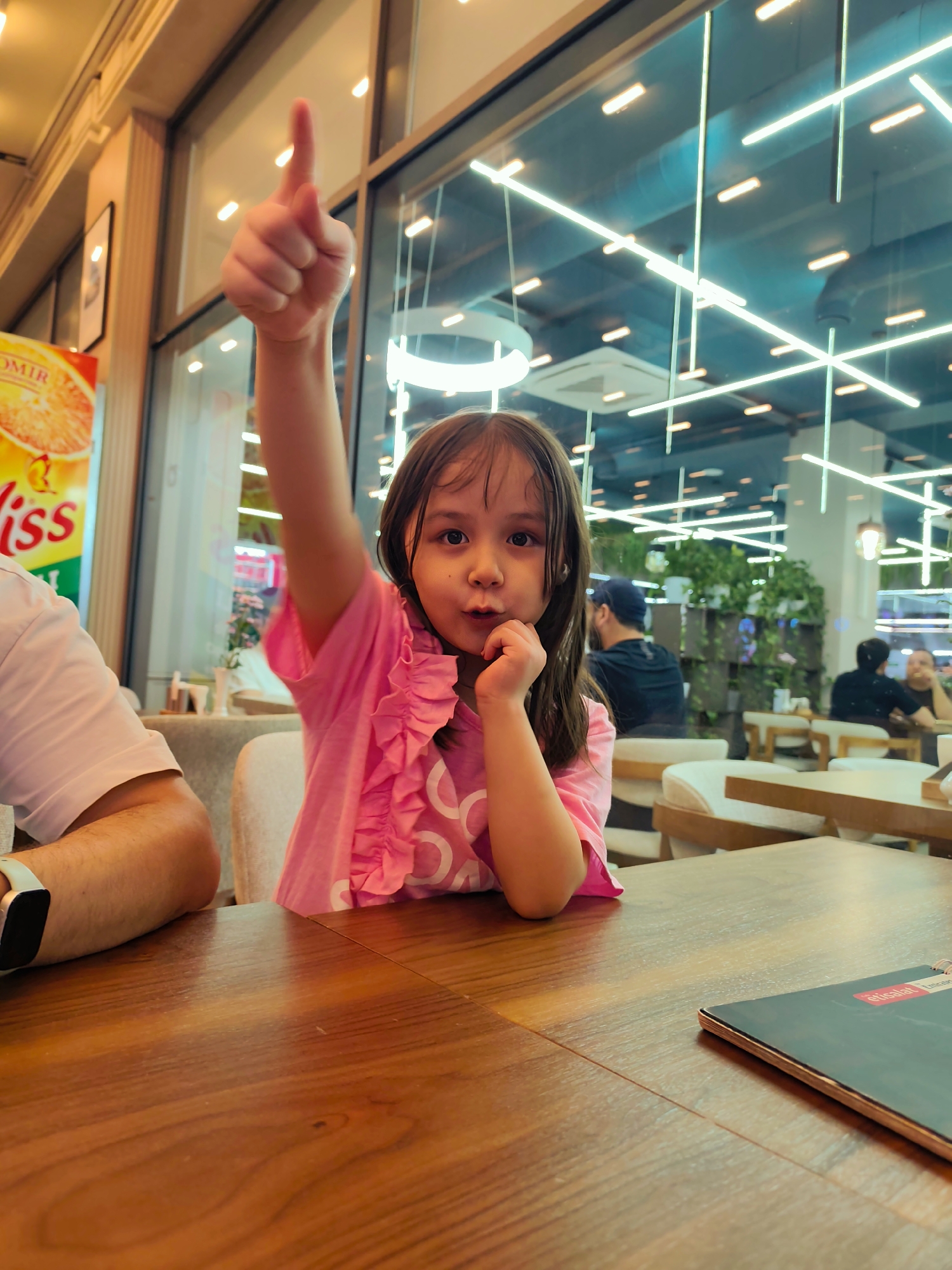A young girl with dark hair, wearing a pink ruffled shirt, sits at a wooden table. She is looking towards the camera with her index finger raised, as if making a point or giving a direction. Her other hand rests on her chin. To her left, part of a person wearing a white shirt and a smartwatch is visible. The background shows a brightly lit interior, possibly a restaurant or cafe, with large windows revealing an outdoor area or another part of the building. Greenery and overhead lights are visible through the windows. The overall scene appears to be indoors, likely during the daytime given the bright lighting. The girl's expression is curious and engaging. A menu is visible on the table in the foreground. Some text is visible on a poster to the left of the girl, reading "OMIR" and "iss". Text on the menu in the foreground is partially obscured but includes "etisalat". The location is identified as Fergana City, Uzbekistan.