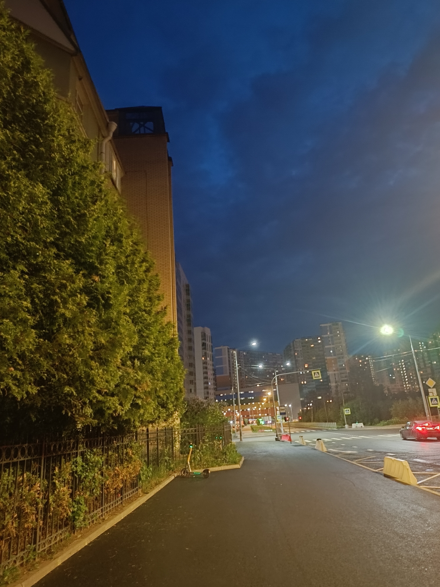 The image depicts a street scene at dusk or night in Saint Petersburg, Russia. The perspective is from a sidewalk looking down a paved road that curves slightly to the right. On the left side of the image, a building with a light-colored brick facade is visible, partially obscured by a dense, green evergreen tree line running along a black metal fence. Beyond the fence, there are some dry bushes and foliage. A lone electric scooter with illuminated green lights is parked on the sidewalk near the fence.
The road is lined with traffic lights, street lamps, and road signs, indicating an urban environment. Several multi-story buildings with illuminated windows are visible in the background, suggesting a residential or commercial area. One of these buildings has a distinctive tower-like structure on top. A traffic signal with red and yellow lights is visible at an intersection further down the road. A red car is turning at the intersection, with its brake lights illuminated. Yellow concrete barriers are placed along the side of the road. The sky is a deep blue with some scattered clouds, and the overall lighting suggests the transition from day to night.