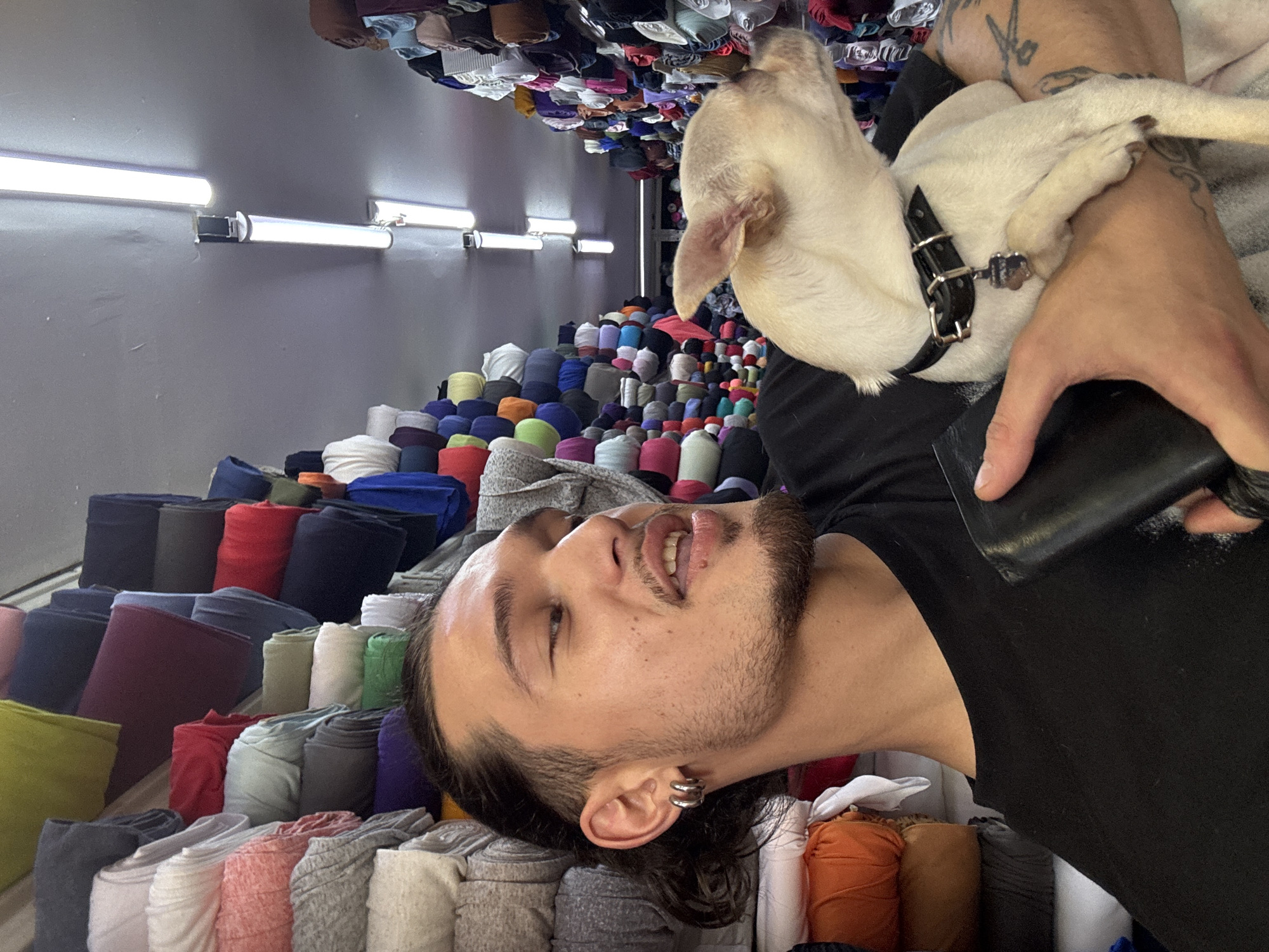 In an indoor textile market, a young man with dark, wavy hair pulled back and two silver hoop earrings in his left ear, is holding a small, light-colored Chihuahua. The man is wearing a black t-shirt and a black wallet is visible in his left hand. The Chihuahua has a black collar with a silver tag. Behind them, aisles are lined with tall stacks of rolled fabrics in various colors, including white, pink, grey, green, blue, red, and orange. Fluorescent lights are visible on the ceiling. This scene is located in the Textile Market, also known as the "Jenite pazar" (Women's Market) in Sofia, Bulgaria.