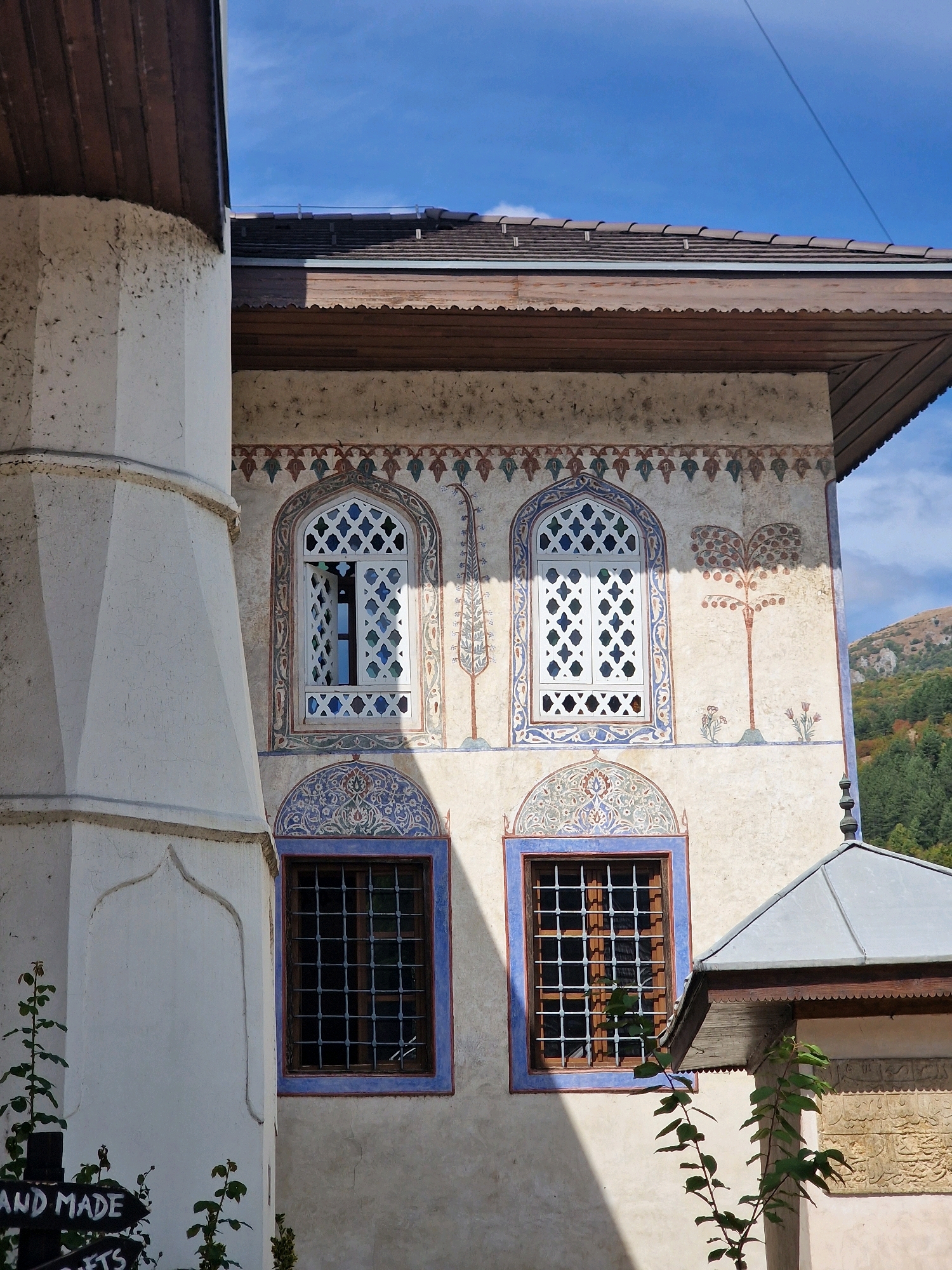 The image shows the exterior of the Šarenica Mosque in Travnik, Bosnia and Herzegovina. The mosque features ornate architectural details, including decorative window frames, colorful frescoes of trees and floral motifs on its facade, and a wooden roof. The building's stucco walls are painted in light colors, contrasting with the dark window grilles and wooden trim. A large, fluted white pillar is visible on the left side of the image, partially obscuring the mosque's facade. In the background, a hillside covered with trees and a clear blue sky with some clouds are visible. The image is taken from a low angle, emphasizing the architectural features of the mosque. Several green plants are growing in the foreground.