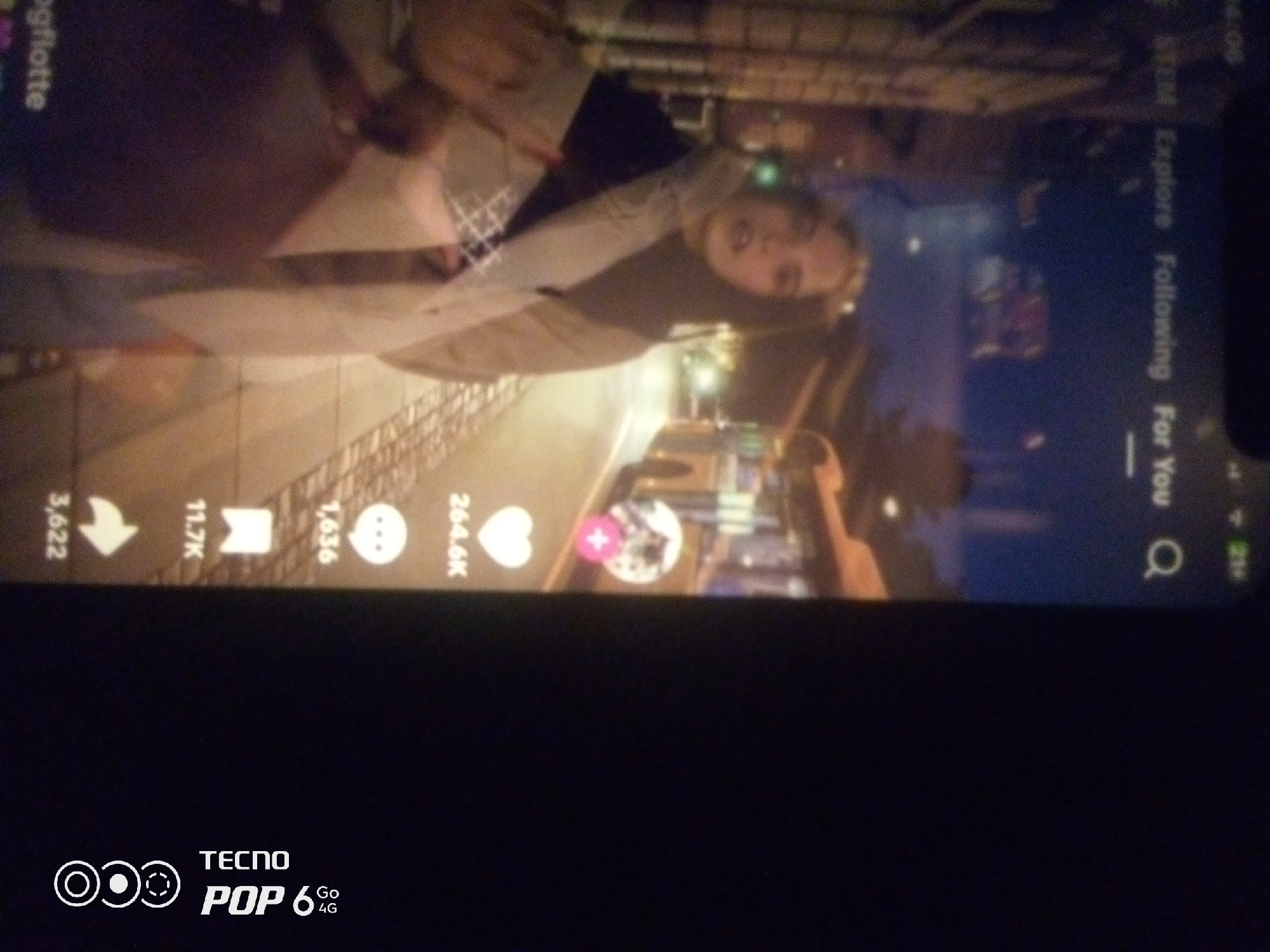 The image depicts a vertical video interface, likely from a social media platform like TikTok, overlaid on a nighttime city scene. The video content features a person, possibly a young woman, with blonde hair, looking towards the camera. Her face is partially obscured by a sheer fabric. The background of the video shows a street with lights and buildings, suggesting an urban environment.
On the right side of the interface, several icons and text are visible, indicating navigation and engagement features: "Explore," "Following," and "For You." Below these, there's a search icon. Along the bottom edge of the video interface, there are numerical counts and icons, likely representing likes, shares, comments, and other forms of interaction. These include "3,622," "11.7K," "1,636," and "264.4K."
The bottom of the image shows the "TECNO POP 6 Go 4G" branding, indicating the device on which the video is being viewed. The overall scene is dark, with the video playing being the primary source of light. The city of Howayi, Nigeria, is not explicitly depicted or mentioned in the visual elements of the image.