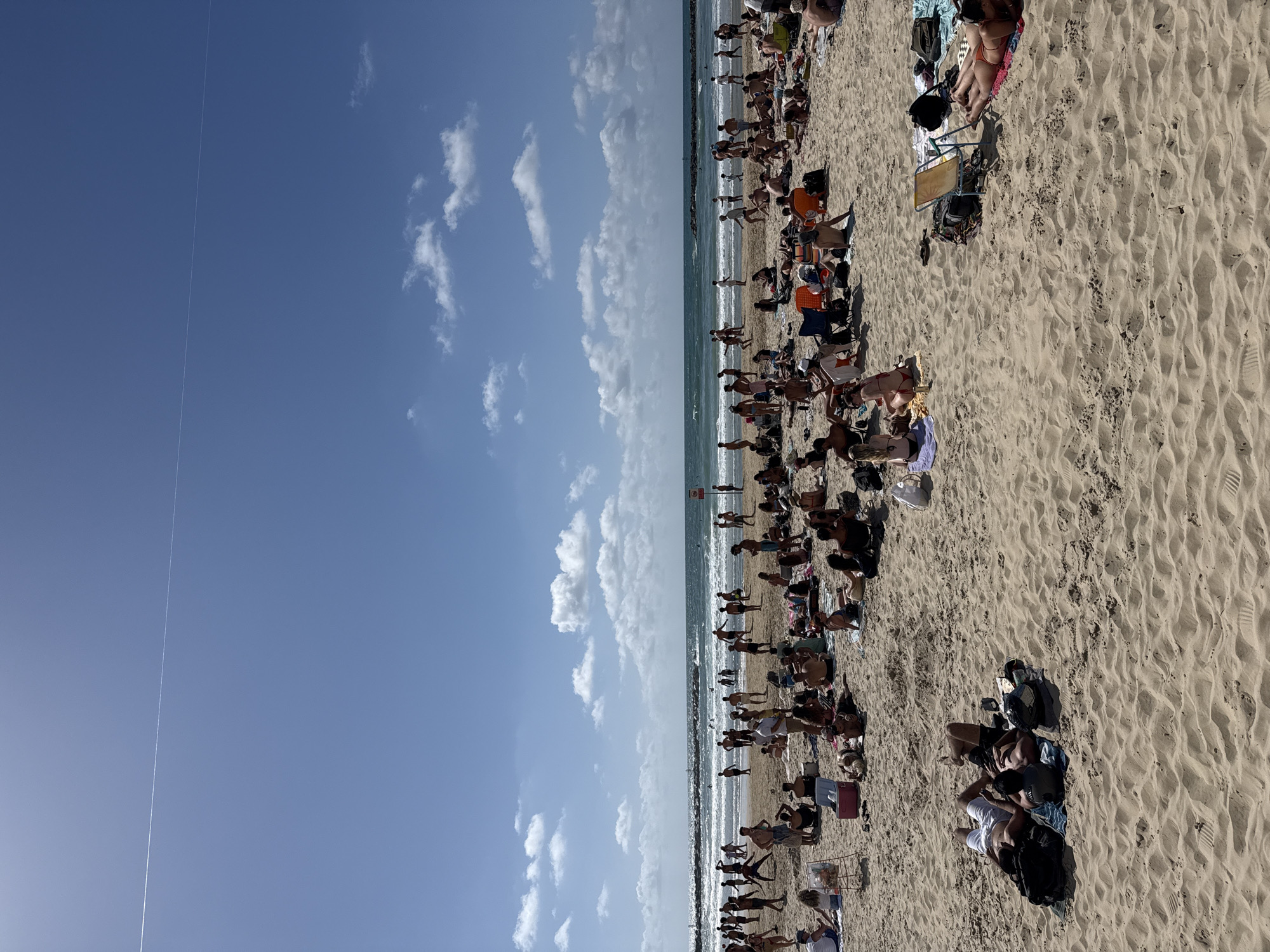 The image depicts a crowded beach scene in Tel Aviv, Israel, on a sunny day. People are lounging on the sand, many are sunbathing, and others are walking or standing near the water's edge. There are numerous beach chairs and personal belongings scattered across the sand. A seawall stretches along the beach and divides the beach. The sky is blue with some scattered white clouds. There is a contrail stretching through the sky.
