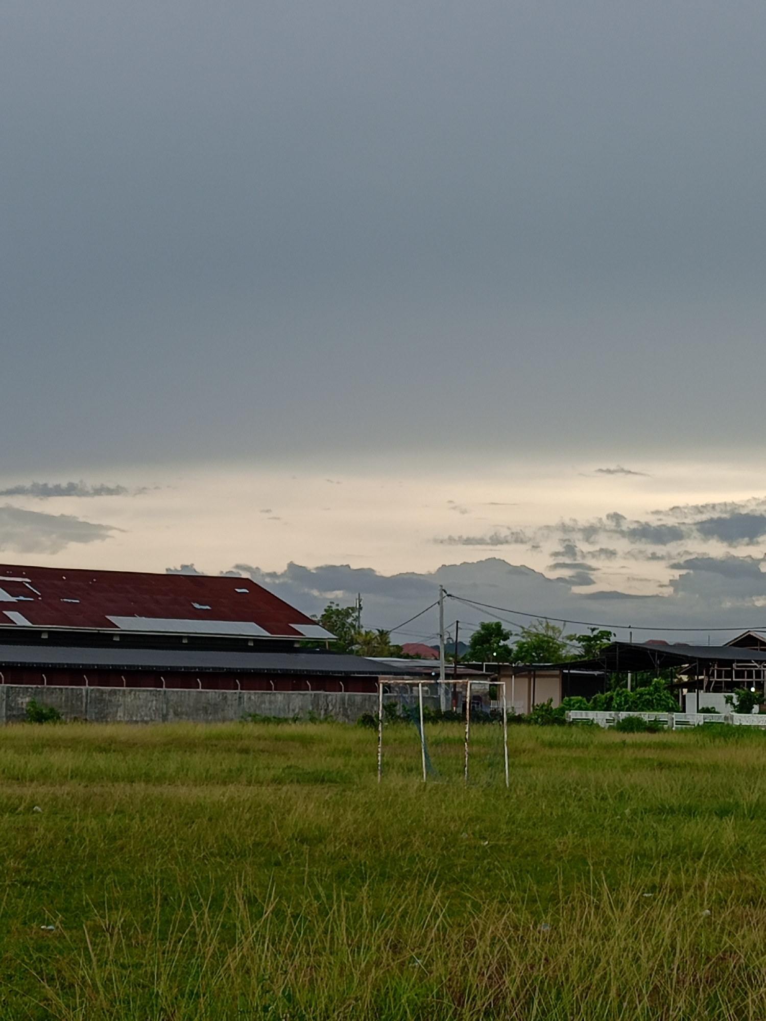 The image shows a grassy field in Geulanggang Baro, Indonesia. In the background, there's a building with a red roof and a row of solar panels. A soccer goal stands in the field. The sky is overcast with dark clouds. Other buildings are visible in the distance beyond the field. Power lines cross the scene.