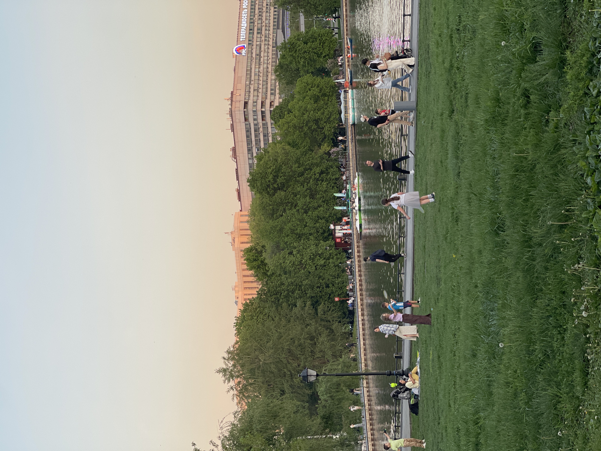 The image shows a park in an unknown city, possibly in Russia given the building with Russian text in the background. A body of water, possibly a pond or a river, runs through the park. People are seen walking along the edge of the water, relaxing on the grass, and playing badminton. Trees and a multi-story building border the park. The sky suggests it is either early morning or late evening. The overall impression is a calm, urban park scene.