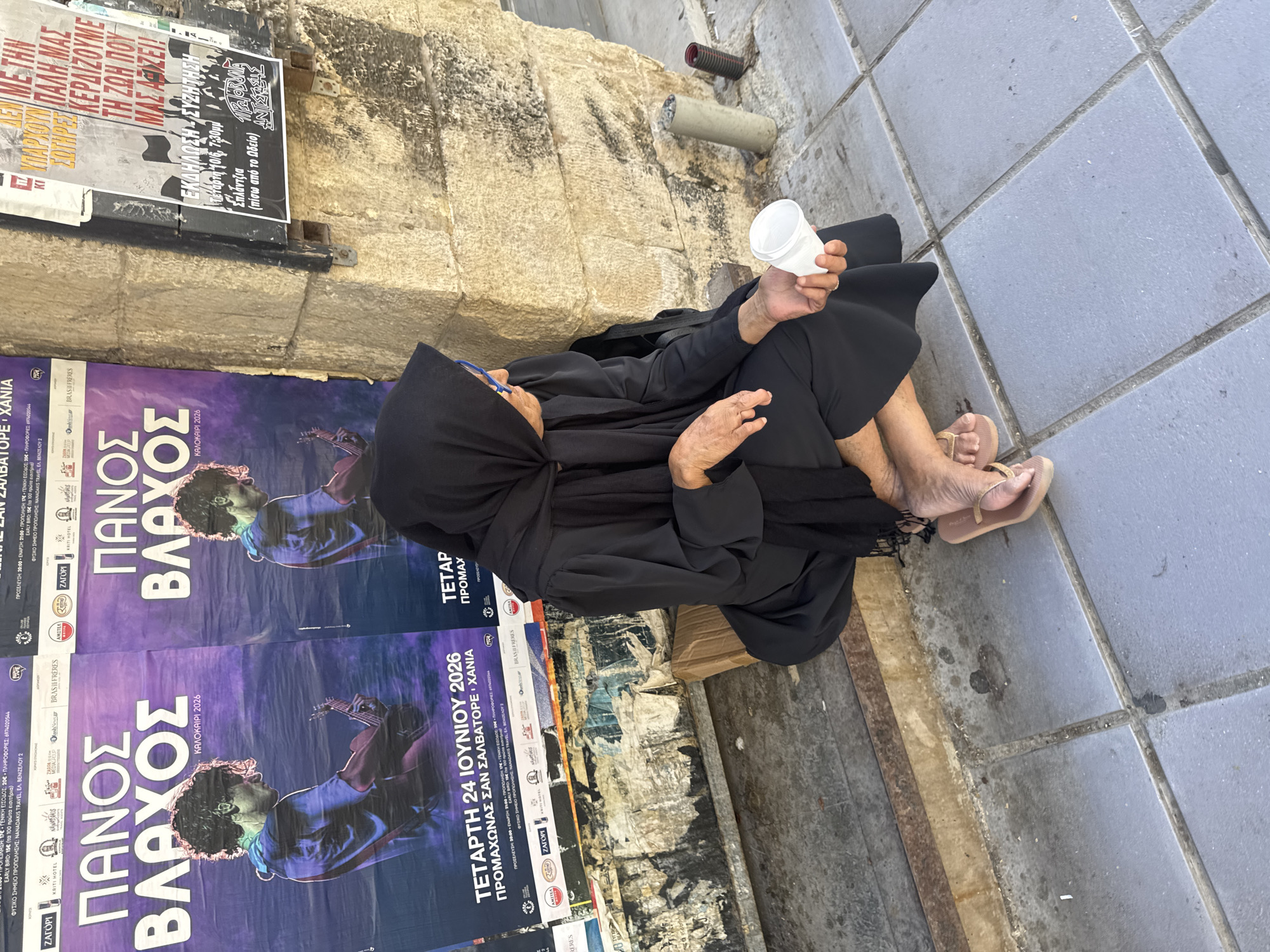 A woman wearing a black hijab and traditional black dress is sitting on a concrete ledge next to a stone building in Chania, Greece. She is holding a white disposable cup in her right hand, appearing to be asking for donations. Behind her are two large posters advertising a performance by Panos Vlachopoulos. The posters are purple with a picture of the performer playing a guitar. To the right of the posters, on the stone wall, are other notices and advertisements. The ground is paved with grey tiles. The scene suggests a public area in the city, possibly near a performance venue or a busy street.