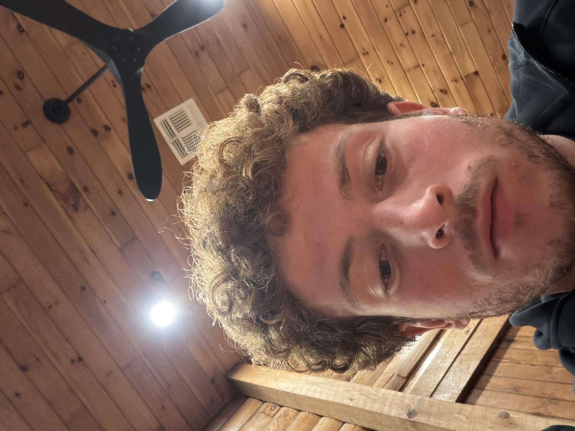 A close-up, angled selfie of a light-skinned male with curly, light brown hair and a short, light brown beard. The subject's head is centered in the frame with a downward gaze. The background consists of a wooden ceiling with visible planks and knots. A black ceiling fan with three blades is located in the upper left corner. A white vent is also visible near the fan. A bright light source is positioned near the center of the background. The subject is wearing a dark garment on the right side of the frame. The image does not contain enough information to determine a specific location in Rock Hill, United States of America.