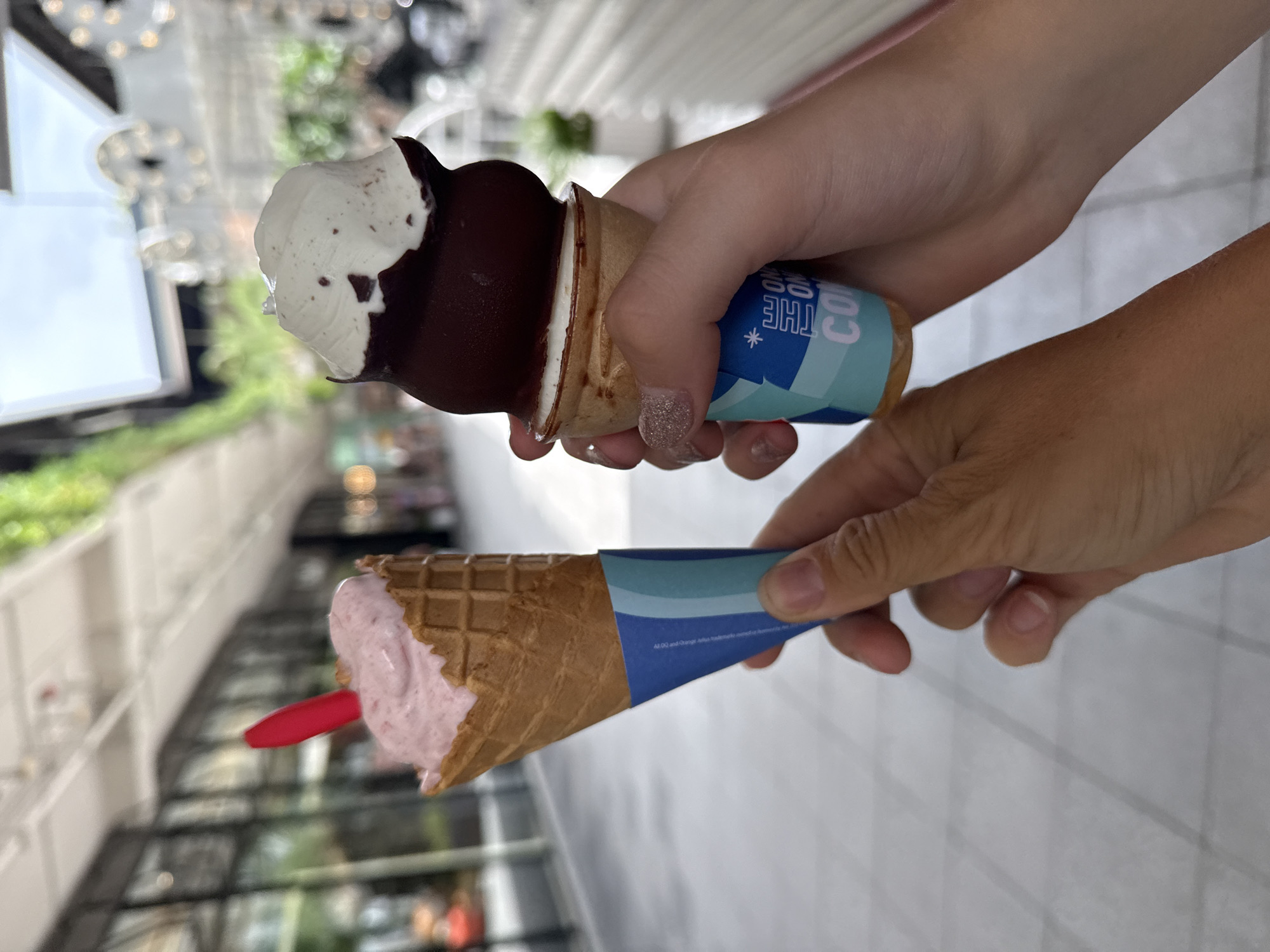 Two hands hold ice cream cones in an outdoor walkway with a blurred background of buildings and greenery. The left hand, holding a strawberry ice cream cone with a red plastic spoon, is to the left and closer to the camera. The ice cream is soft-serve and sits in a waffle cone with a blue and white striped paper wrapper. The right hand, holding a chocolate-dipped ice cream cone topped with white cream, is to the right and slightly behind the left hand. This cone also has a blue and white patterned paper wrapper with text that reads "THE ONE CONE". The hands and ice cream are in focus, while the background is out of focus. The ground appears to be paved with light-colored tiles. The scene suggests a casual outing in a public space, possibly a commercial or tourist area in the city of Ko Samui District, Thailand.