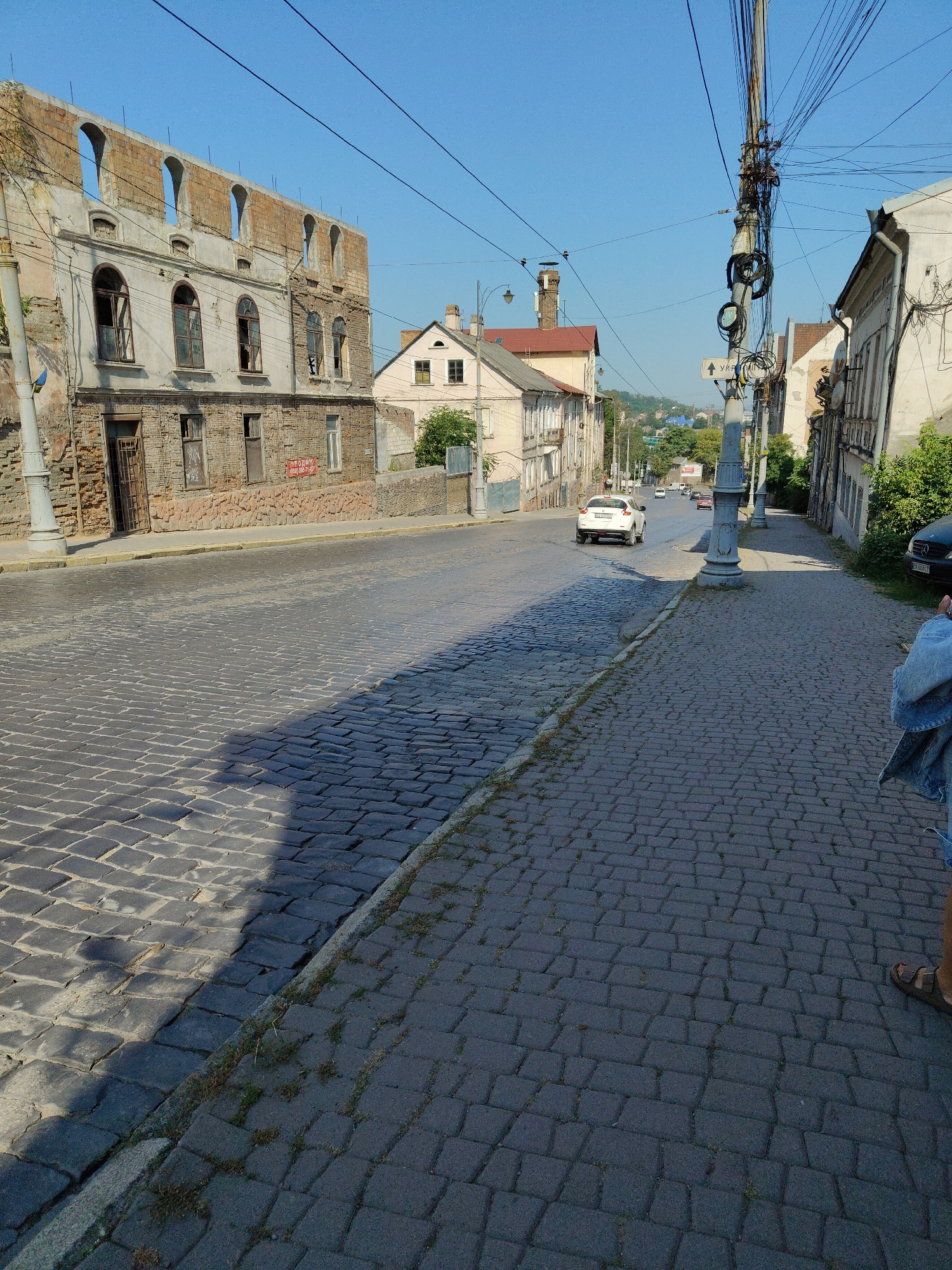 The image depicts a street scene in Chernivtsi, Ukraine, characterized by its cobblestone pavement and older architecture. In the foreground, a paved sidewalk runs parallel to the street. The street itself is paved with dark, irregular cobblestones, and a distinct shadow from an unseen object stretches across it, indicating sunlight from the right.
On the left side of the street, a dilapidated building stands, its upper portion partially dismantled, revealing brickwork and arched openings. The lower sections of this building show windows and a metal grate. Further down the street, other buildings line the road, some appearing older with multi-story facades and arched windows, while others are lighter in color and appear more residential or commercial.
The street is active with traffic. A white car is visible driving away from the viewer on the right side of the street, and other vehicles can be seen further in the distance. A prominent utility pole with a dense tangle of wires stands on the right side of the street, alongside a lamppost.
The background reveals more of the city's urban landscape, with trees and buildings extending towards a hill. The sky is clear and blue. On the right side of the image, the lower legs and feet of a person wearing denim shorts and sandals are visible, suggesting the photograph was taken by someone standing on the sidewalk. A dark sedan is parked on the far right. One of the buildings on the left has a red sign with Cyrillic text, which appears to be an advertisement or notice. The overall impression is of a historic urban environment undergoing gradual change or decay, under a bright, sunny sky.