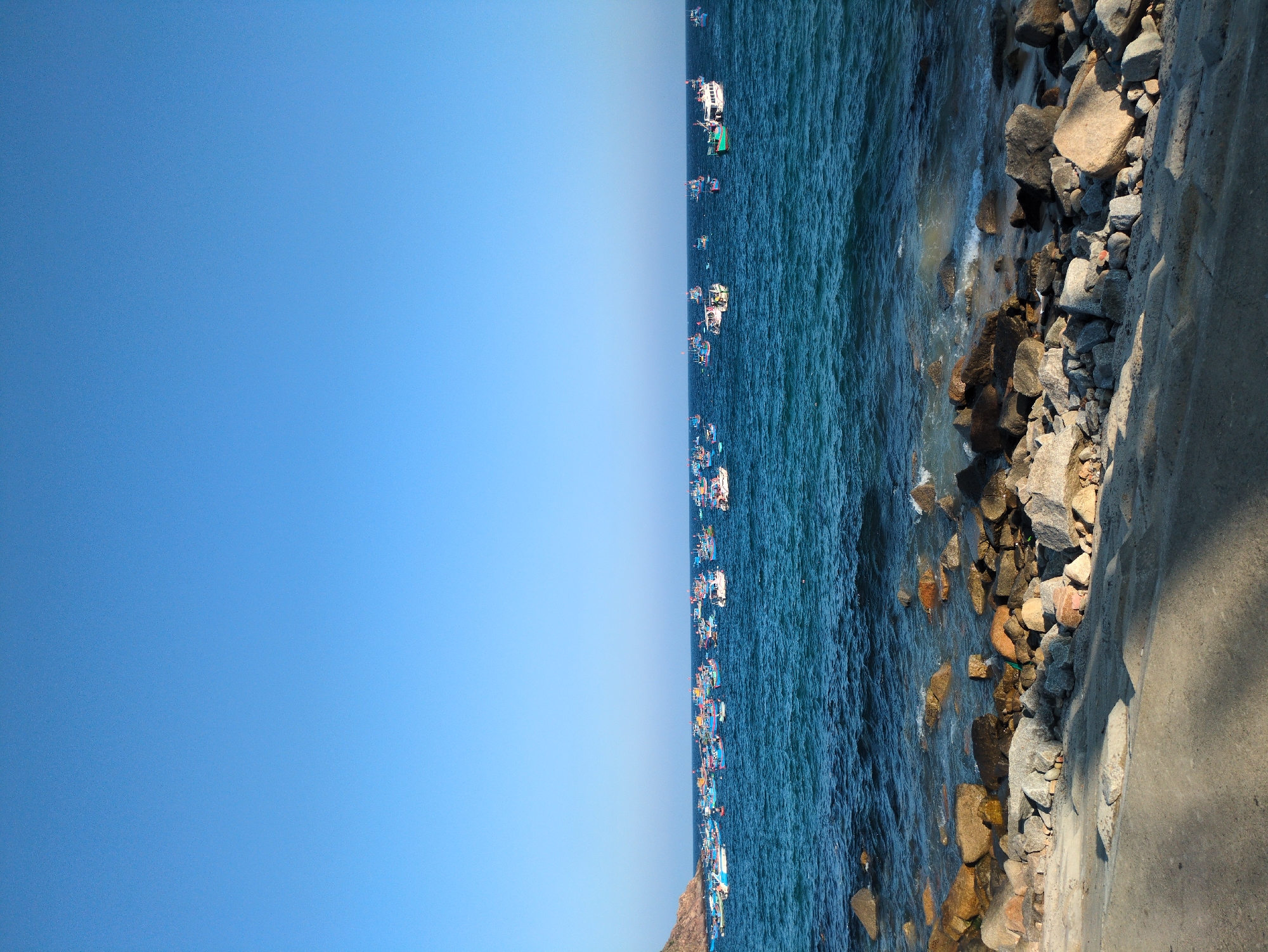 The image captures a coastal scene in East Quy Nhon Ward, Vietnam, under a clear, bright blue sky that occupies the upper third of the frame. The mid-ground is dominated by deep blue-green ocean water, upon which a large cluster of various fishing boats is visible. These vessels are diverse in size and color, featuring traditional Vietnamese fishing boats and larger white motorboats, painted predominantly in blue, red, green, and yellow. A dark, rocky landmass, likely a headland, is present on the far left horizon, partially obscured by the boats. The immediate foreground consists of a rocky shoreline where small waves gently break against a natural arrangement of light grey, beige, and dark brown boulders and smaller stones. A textured, grey concrete path or embankment runs horizontally across the bottom of the frame, partially illuminated by sunlight and cast with shadows. No individuals, specific actions, or interactions are discernible within the frame.