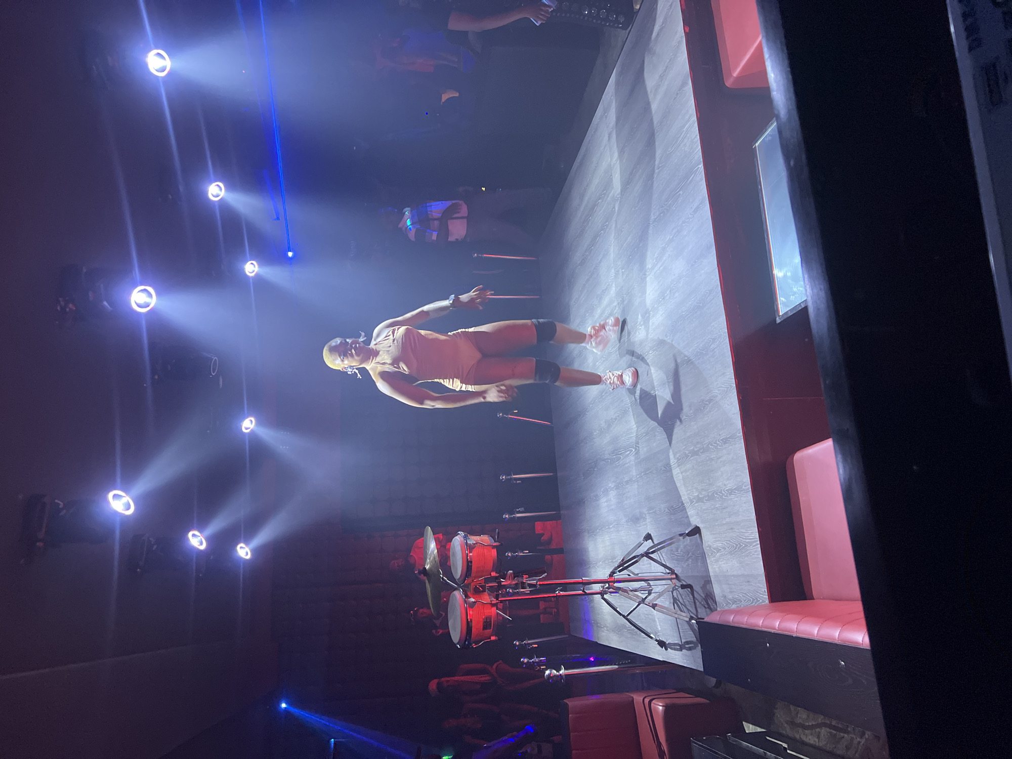 A full shot shows a woman performing on stage in a dimly lit club. The woman is wearing a skin-toned leotard and white sneakers. She has on black knee pads and her hair is short and blonde. She is dancing with her arms outstretched and her body angled to the side. The stage is made of wood and is lit with spotlights from above. To the left of the stage, a set of bongos on a stand are visible. In the background, other club-goers can be seen, some seated in red booths. The overall ambiance of the scene suggests a live performance in a vibrant nightlife setting. The image does not provide any specific details to determine the city of Howayi, Nigeria.