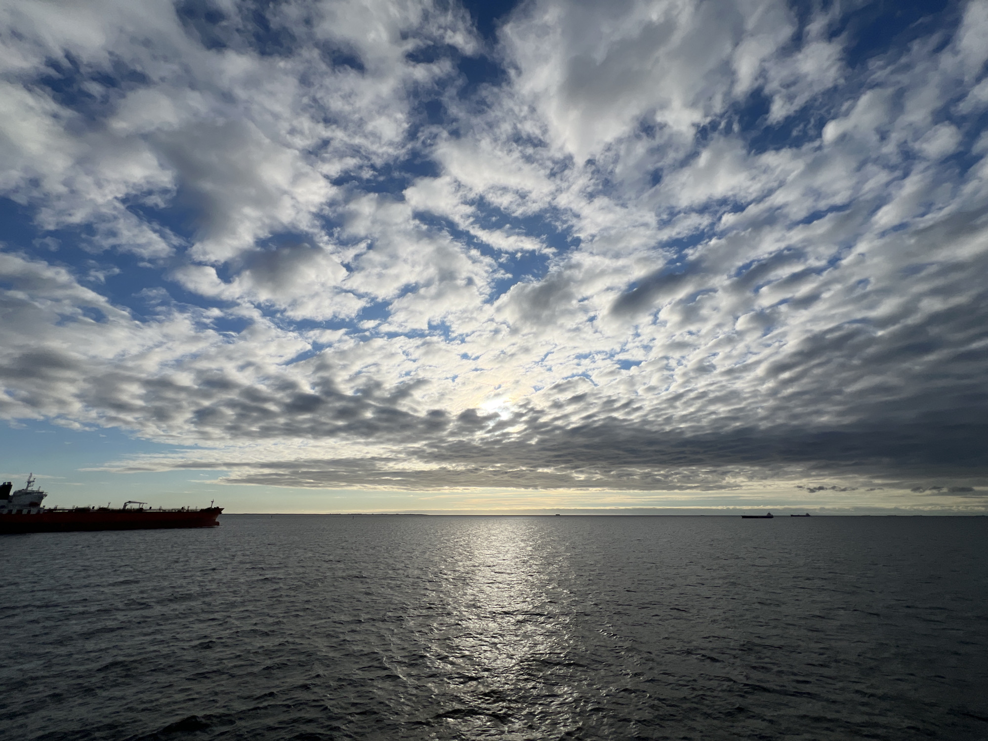 The image depicts a wide expanse of the sea under a partly cloudy sky. The sun is partially obscured by clouds, casting a bright reflection on the water's surface. A large, red cargo ship is visible on the left side of the frame, sailing on the dark blue water. In the distance, several other ships are faintly visible on the horizon. The sky is filled with a dramatic display of clouds, ranging from bright white to dark gray, creating a dynamic contrast with the blue sky peeking through. The overall scene conveys a sense of maritime activity and the vastness of the ocean. The location is the sea off the coast of Gdynia, Poland, likely within the Baltic Sea.