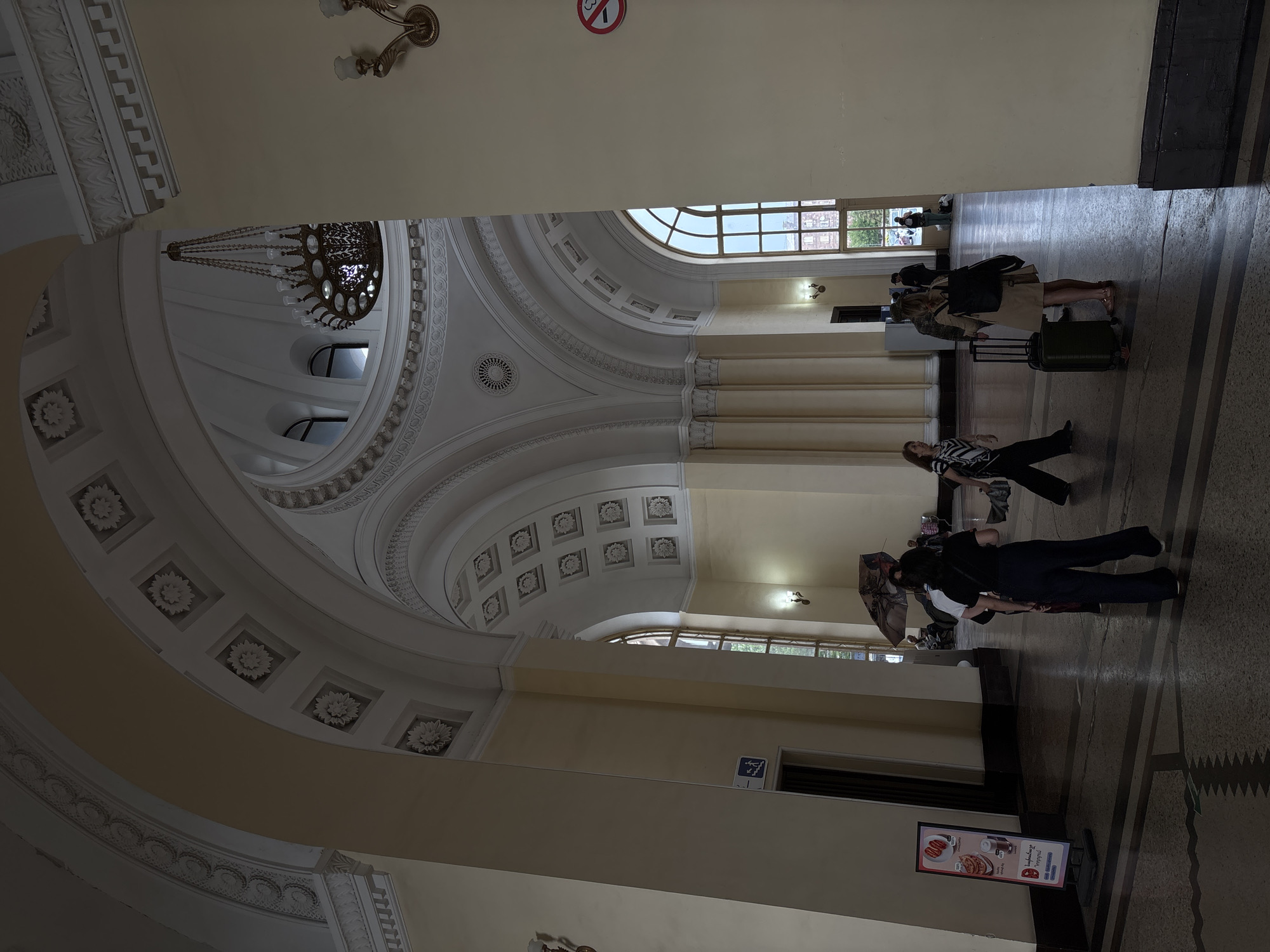 The image depicts the interior of a grand, classical-style building, likely a train station, with high vaulted ceilings adorned with decorative moldings and circular motifs. A large, ornate chandelier hangs from the central dome. Sunlight streams through tall, arched windows, illuminating the space. Several people are present, some walking with luggage, suggesting travel. One individual is holding an umbrella. A sign with food imagery and text in an unfamiliar language is visible on the left. The architecture suggests a location with historical significance. Based on visual cues, this scene is captured at the Yerevan Railway Station in Yerevan, Armenia.